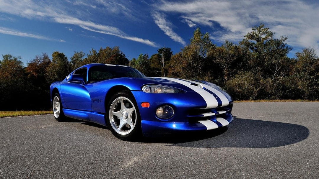Blue Dodge Viper GTS with white racing stripes parked on asphalt with a forest and blue sky background.
