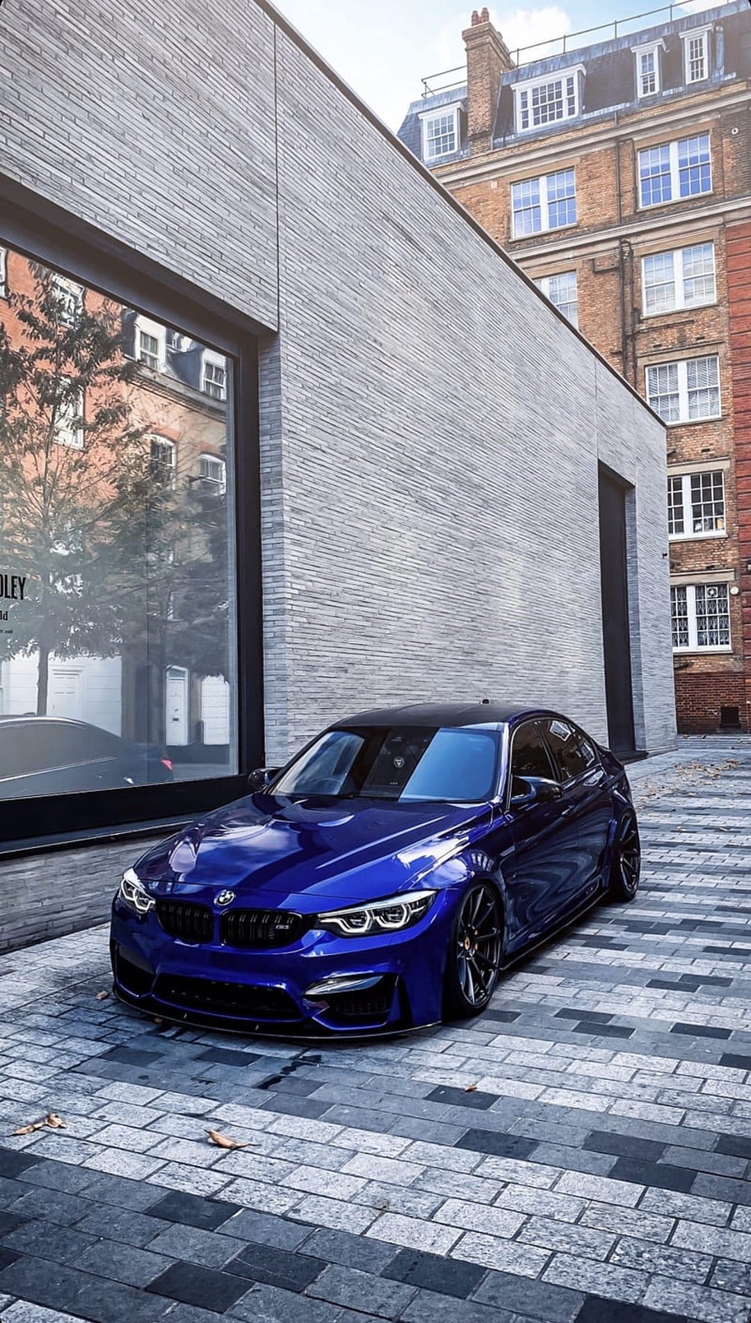 Blue BMW M4 with an aggressive black grille parked on a cobblestone street in front of a modern glass building.