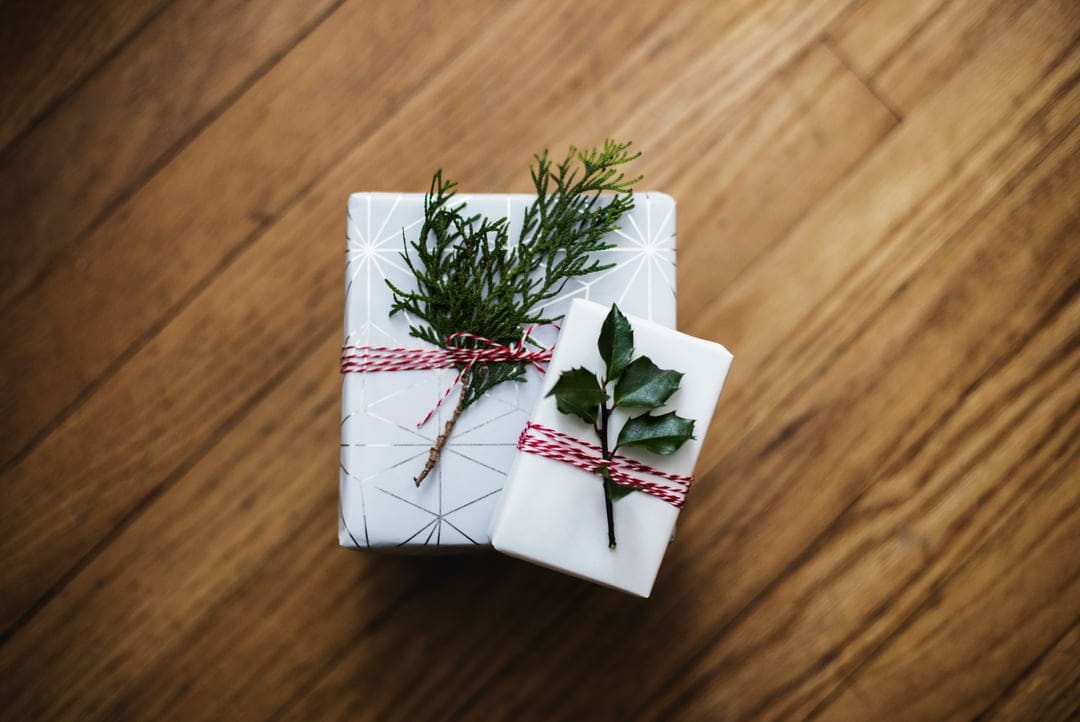 Two white wrapped gifts with evergreen sprigs and red twine sit on a warm brown wood floor from an overhead view.