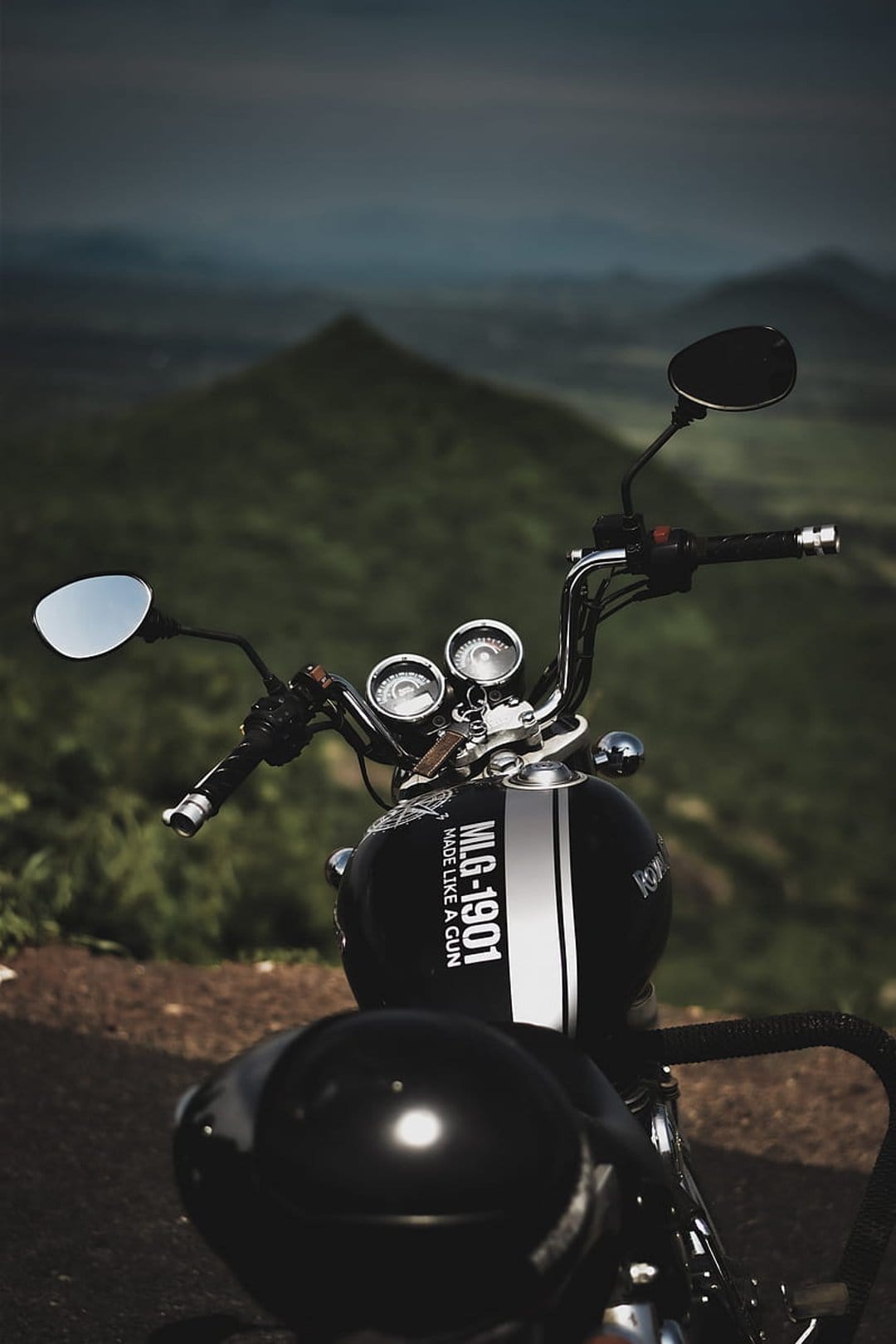 Vintage motorcycle handlebars and a striped fuel tank face a lush green mountain range on a rugged dirt path.