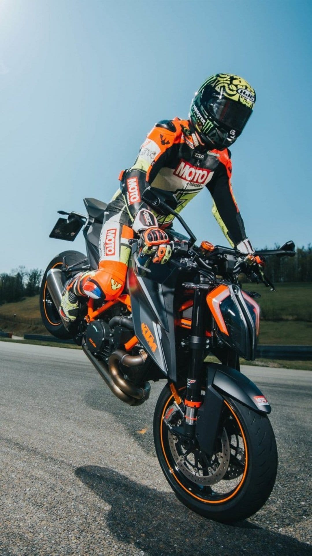 KTM rider in orange and black racing gear performs a motorcycle wheelie against a bright blue sky and green hills.