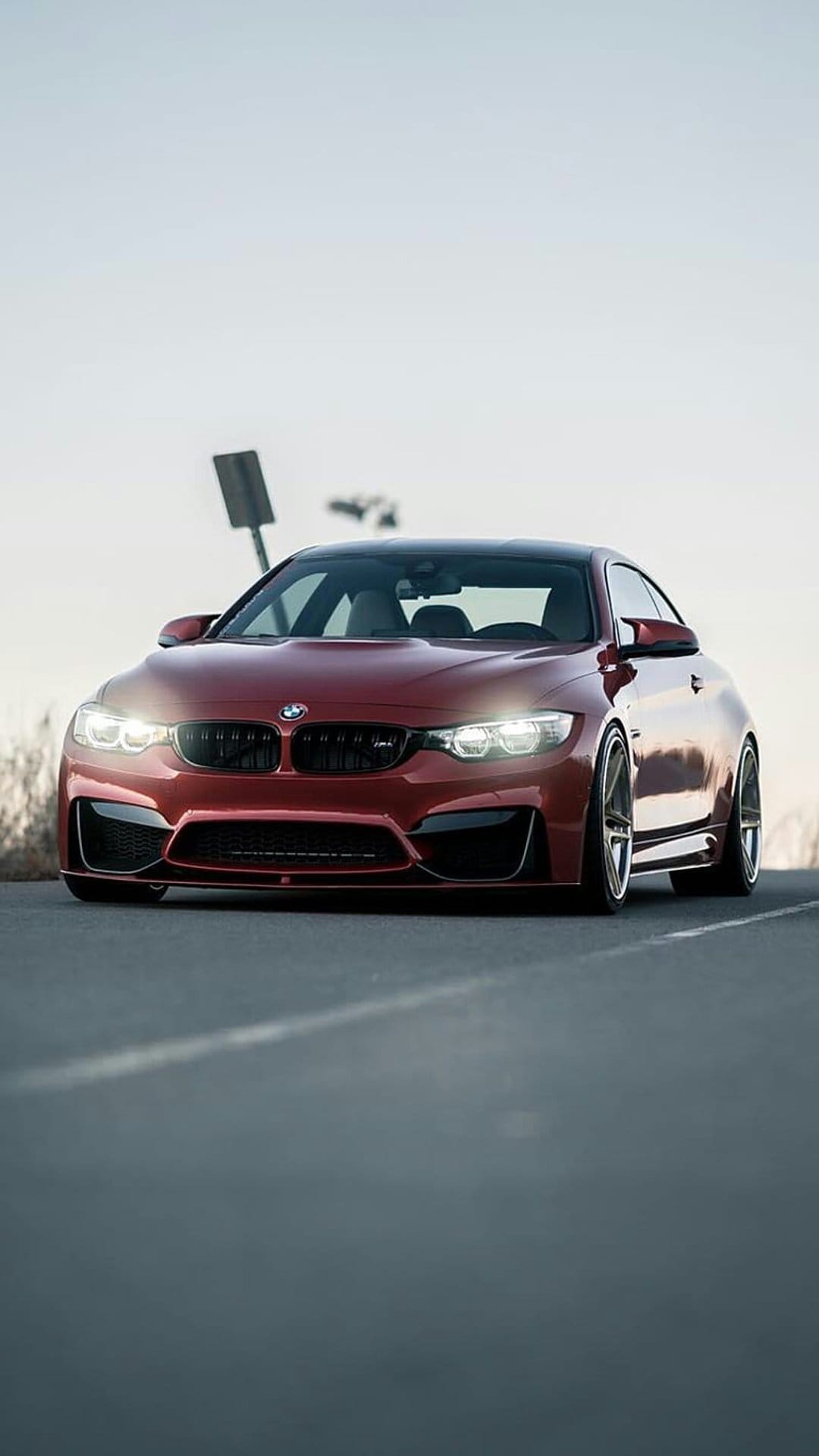 Red BMW M4 Coupe with glowing headlights drives on a highway road at sunset against a bokeh horizon backdrop.