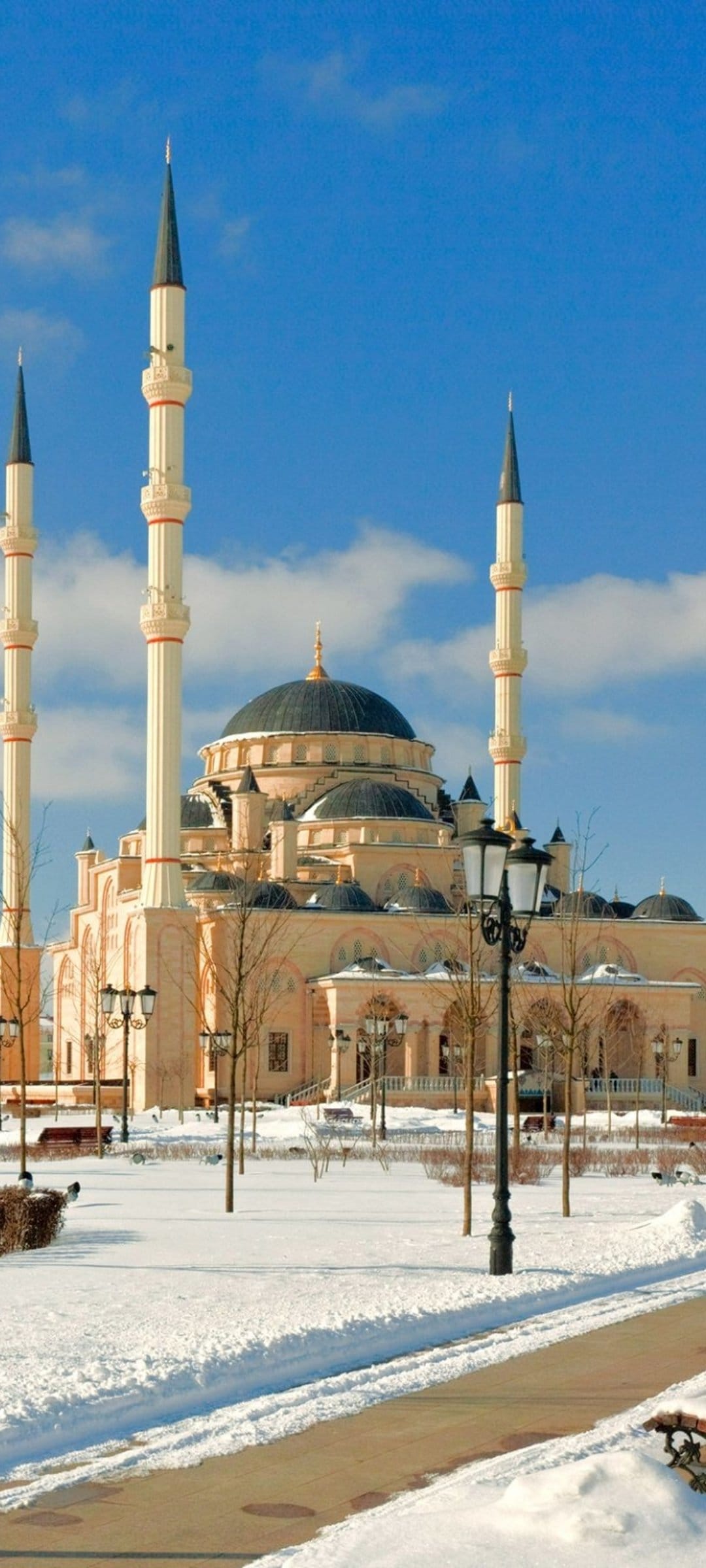 Cream-colored mosque with tall minarets stands in a snowy courtyard under a blue sky with scattered white clouds.