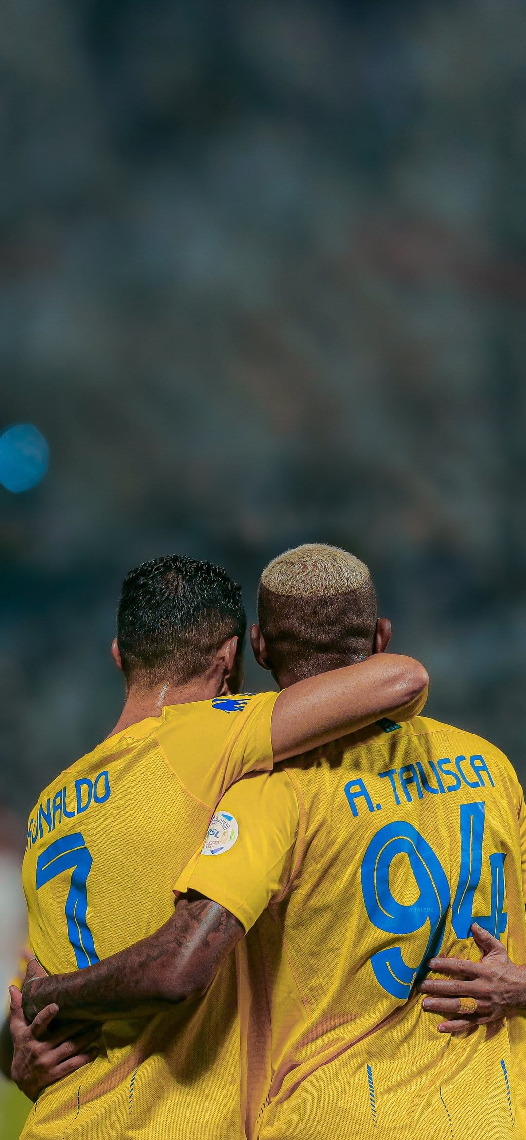 Cristiano Ronaldo and Anderson Talisca embrace in yellow Al Nassr jerseys on a blurred green football pitch.