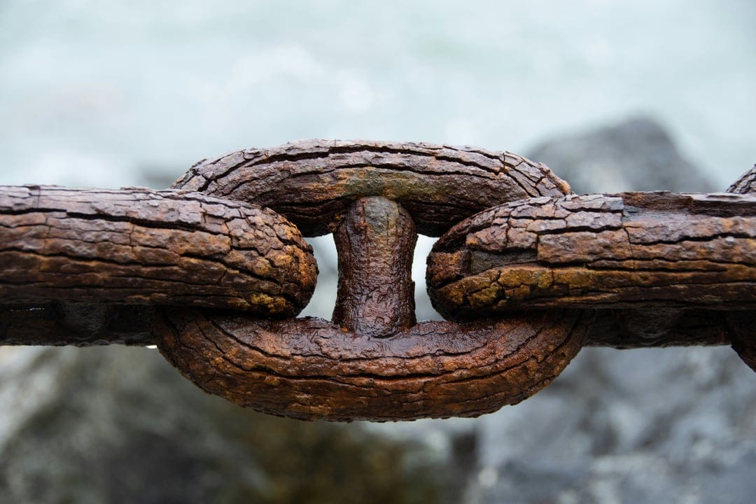 Thick rusted iron chain links with a textured orange patina over a bokeh background of grey rocks and water.