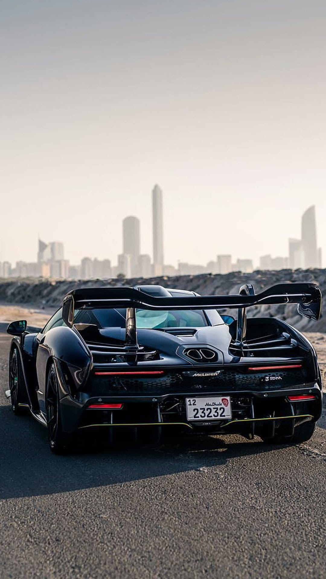 Black McLaren Senna supercar rear view featuring a massive wing and LED lights with a blurred city in the background.