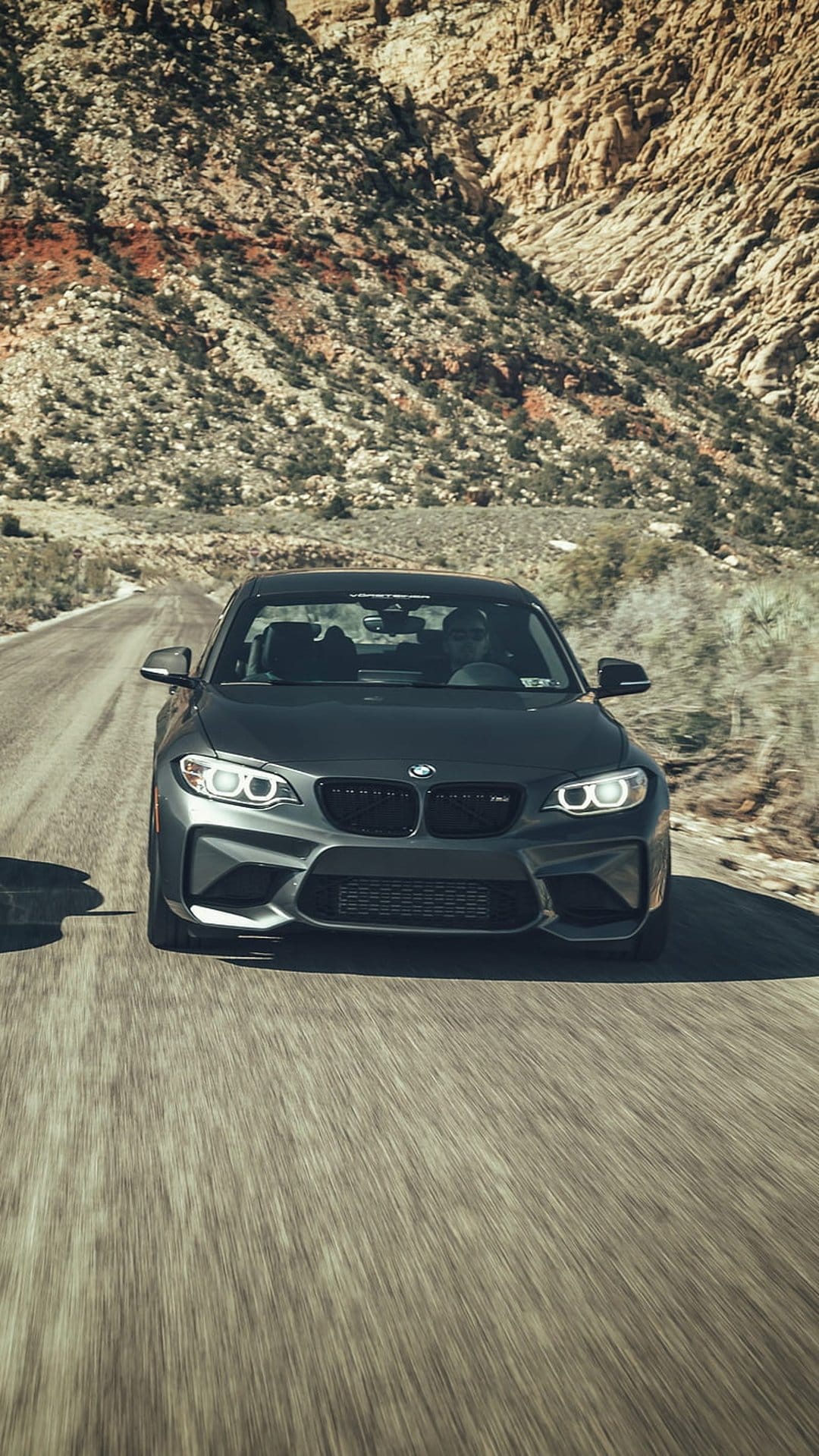 BMW M2 sports car driving on a paved desert road with rugged mountains and motion blur in the background.