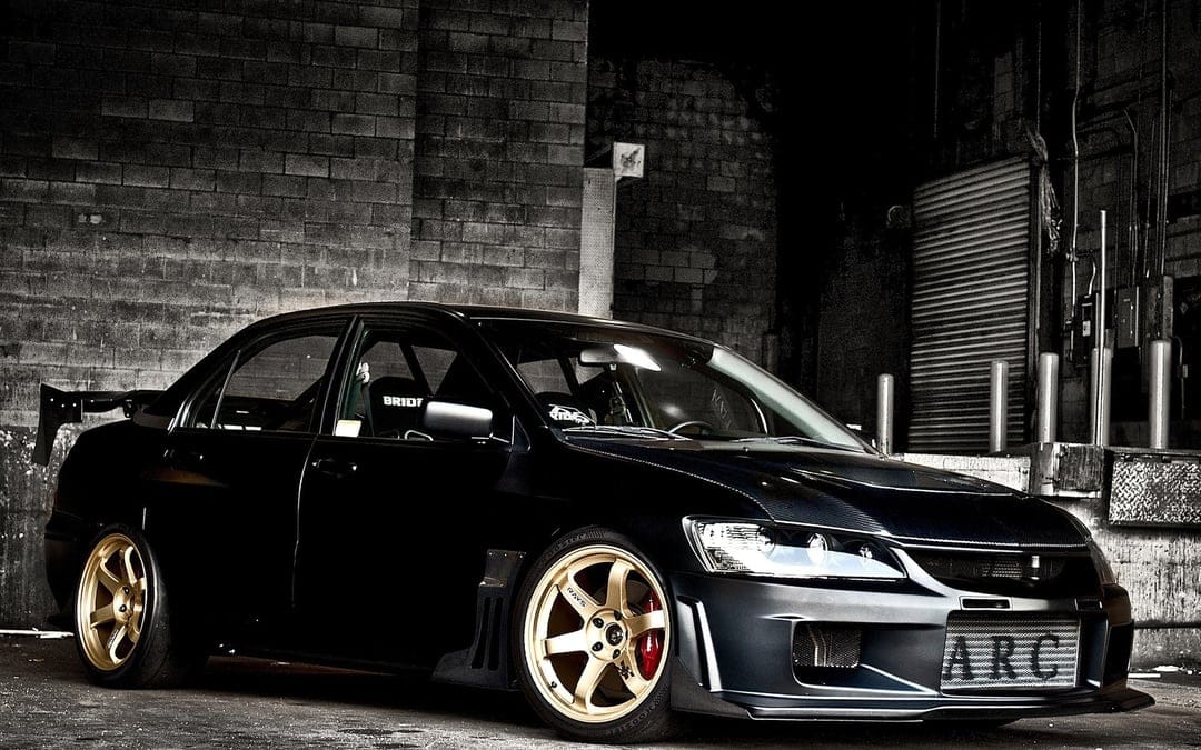 Black Mitsubishi Lancer Evolution with gold wheels and carbon fiber hood in a dark industrial garage.