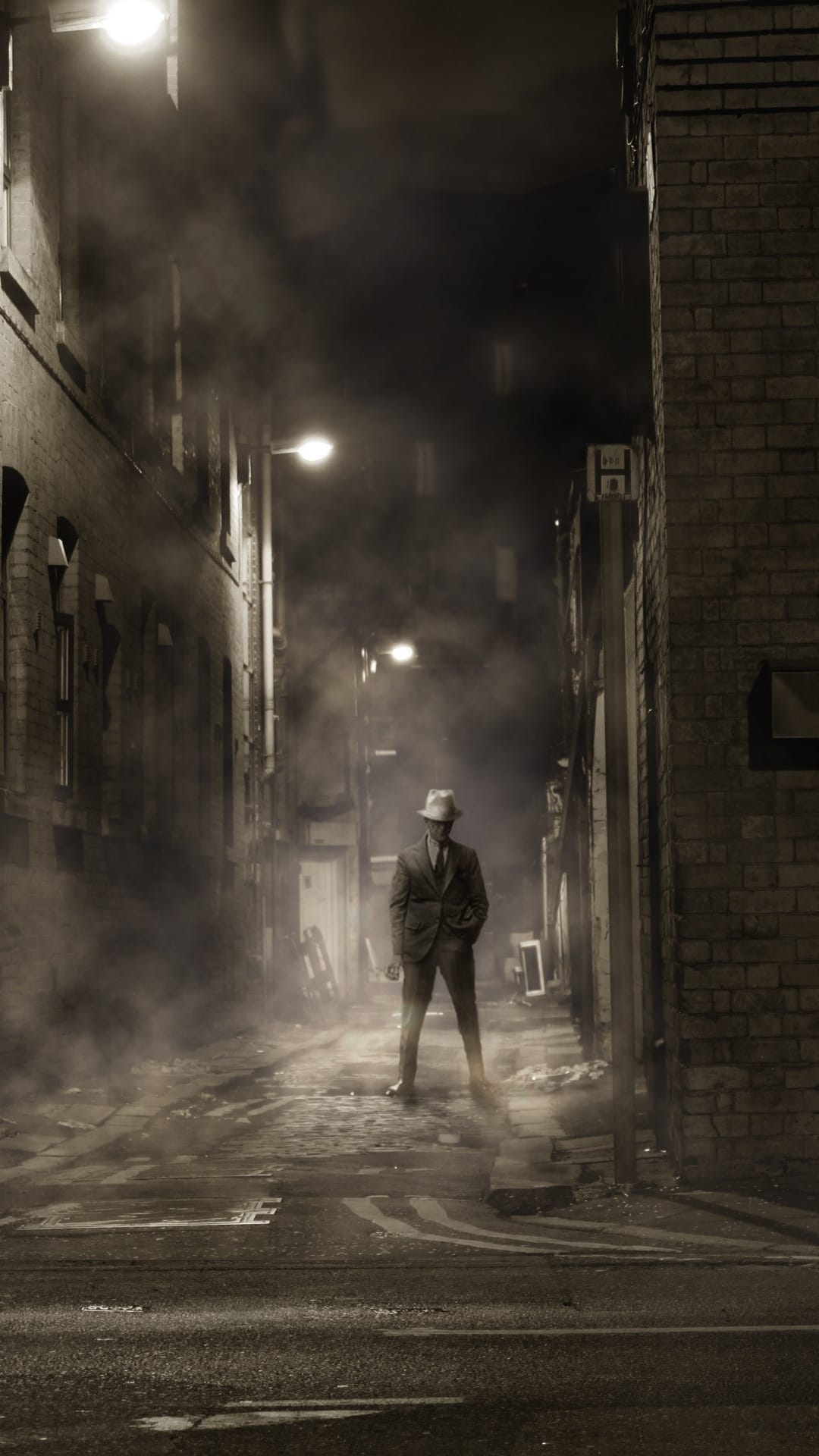 Man in a fedora and suit standing in a dark, misty brick alleyway under dim yellow streetlights at night.
