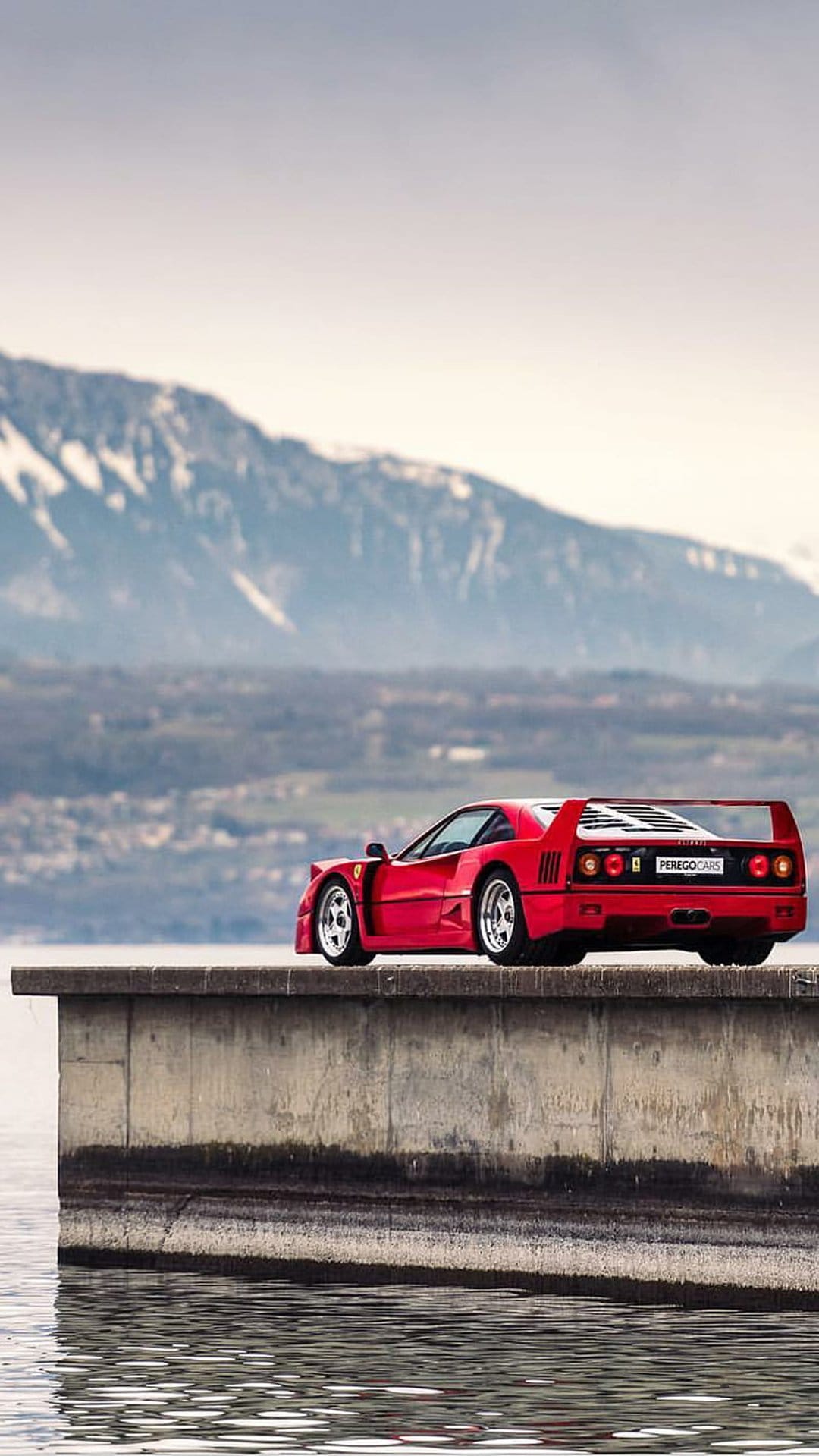 Red Ferrari F40 sports car parked on a concrete ledge by calm water with snow-capped mountains in the background.