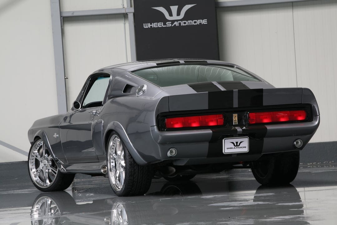 Rear view of a gray classic Shelby GT500 muscle car with black racing stripes and polished chrome rims in a studio.