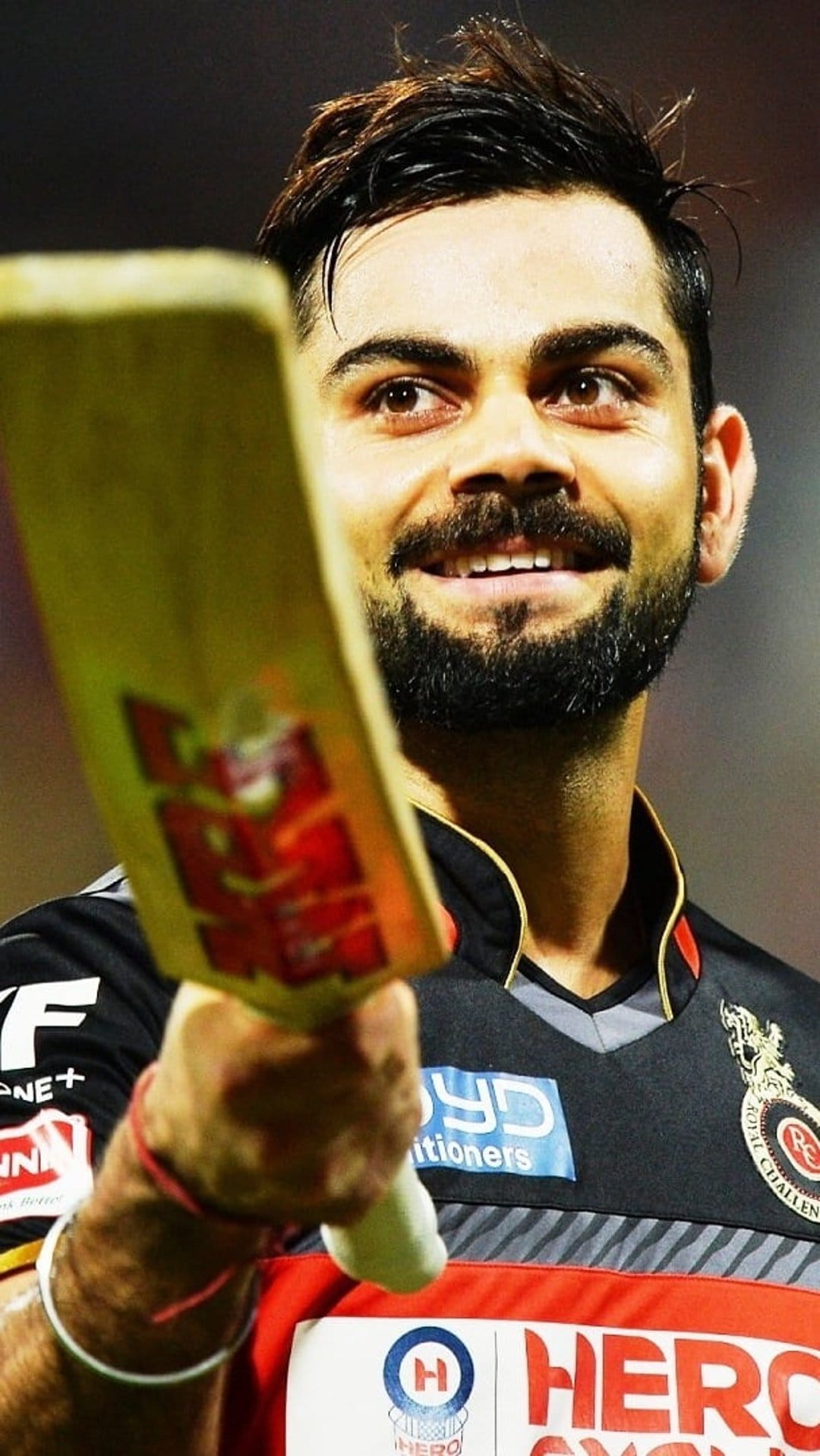 Virat Kohli smiling and holding a cricket bat while wearing a red and gold Royal Challengers Bangalore jersey.