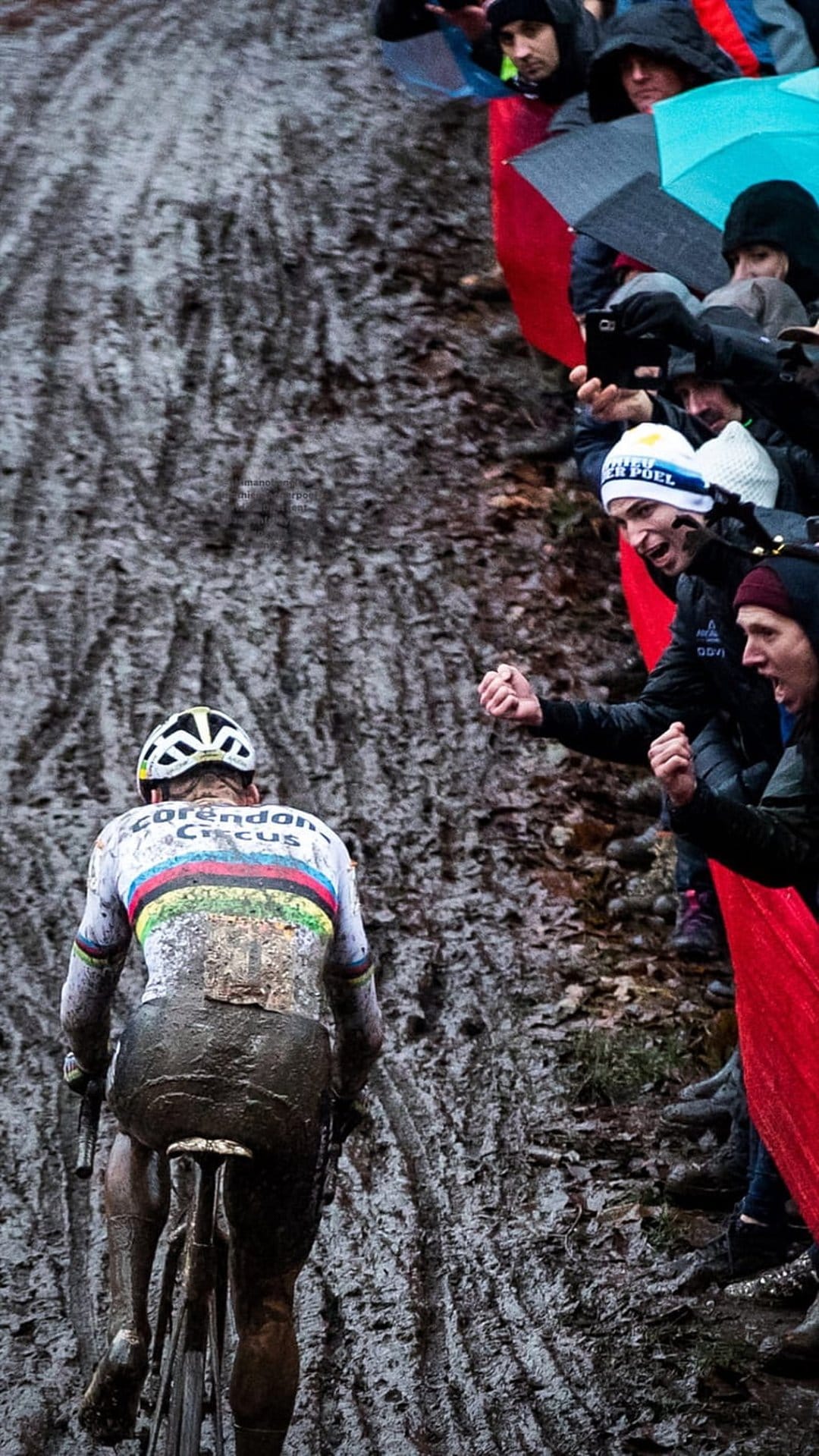 Mud-splattered cyclocross racer navigates a wet track while spectators hold umbrellas and flags in the background.