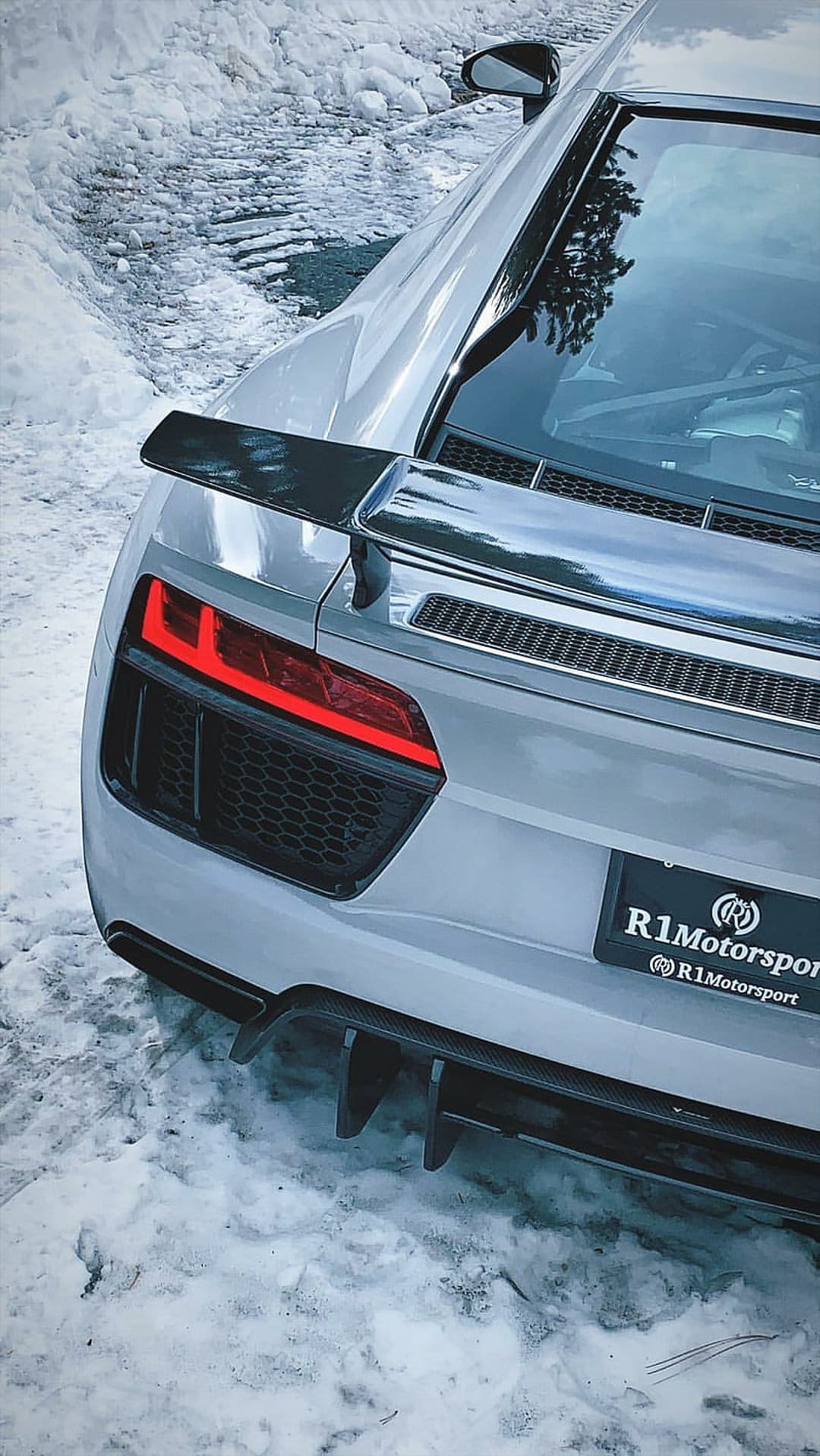Silver Audi R8 supercar rear with a large spoiler and red taillights parked in a textured white snow field.