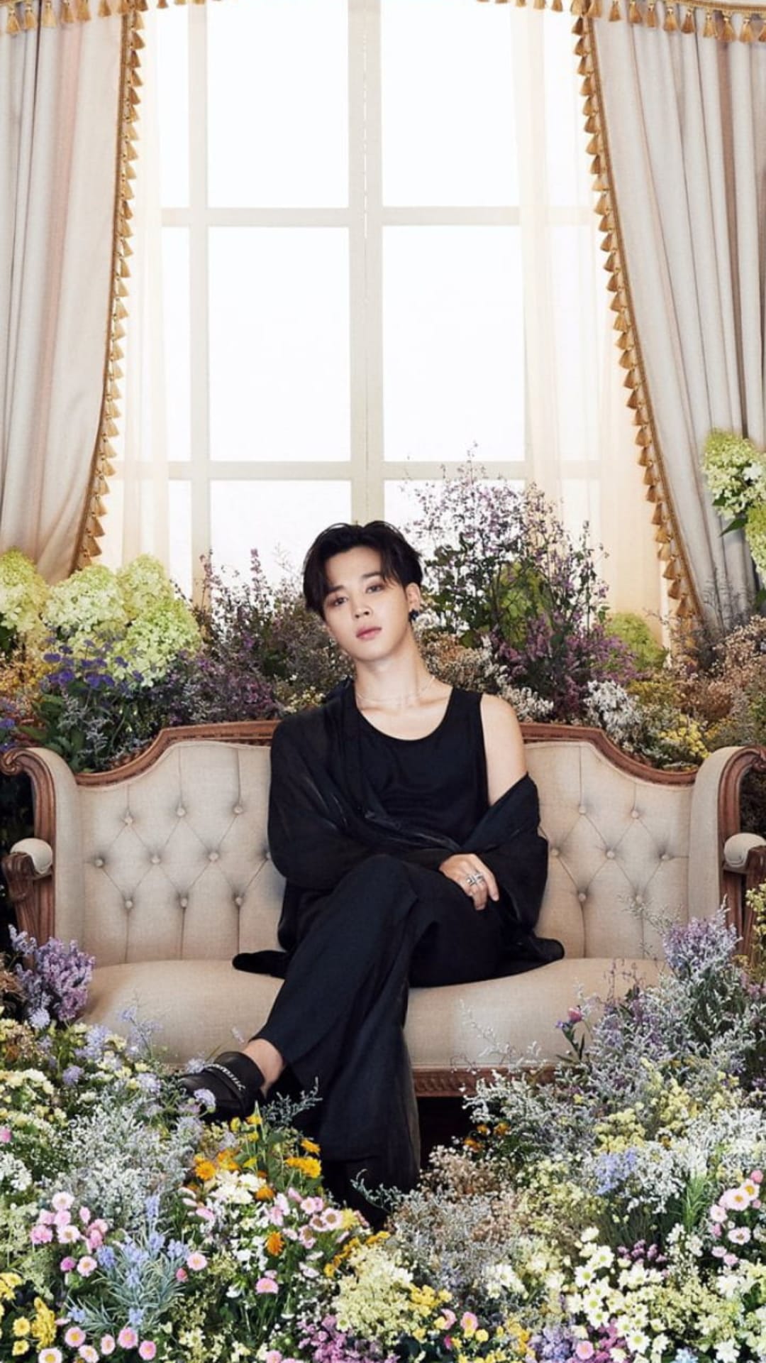 Park Jimin sits on a vintage couch surrounded by colorful flowers while wearing a stylish black sleeveless outfit.