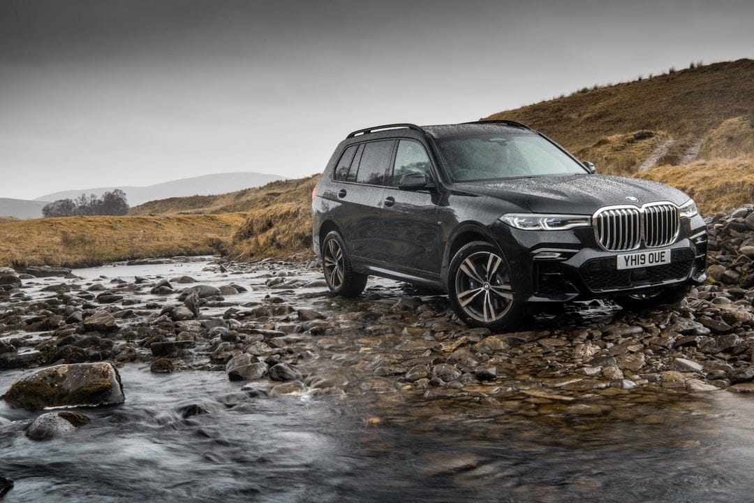 Black BMW SUV parked on smooth river rocks within a flowing stream under a gray overcast sky during light rain.
