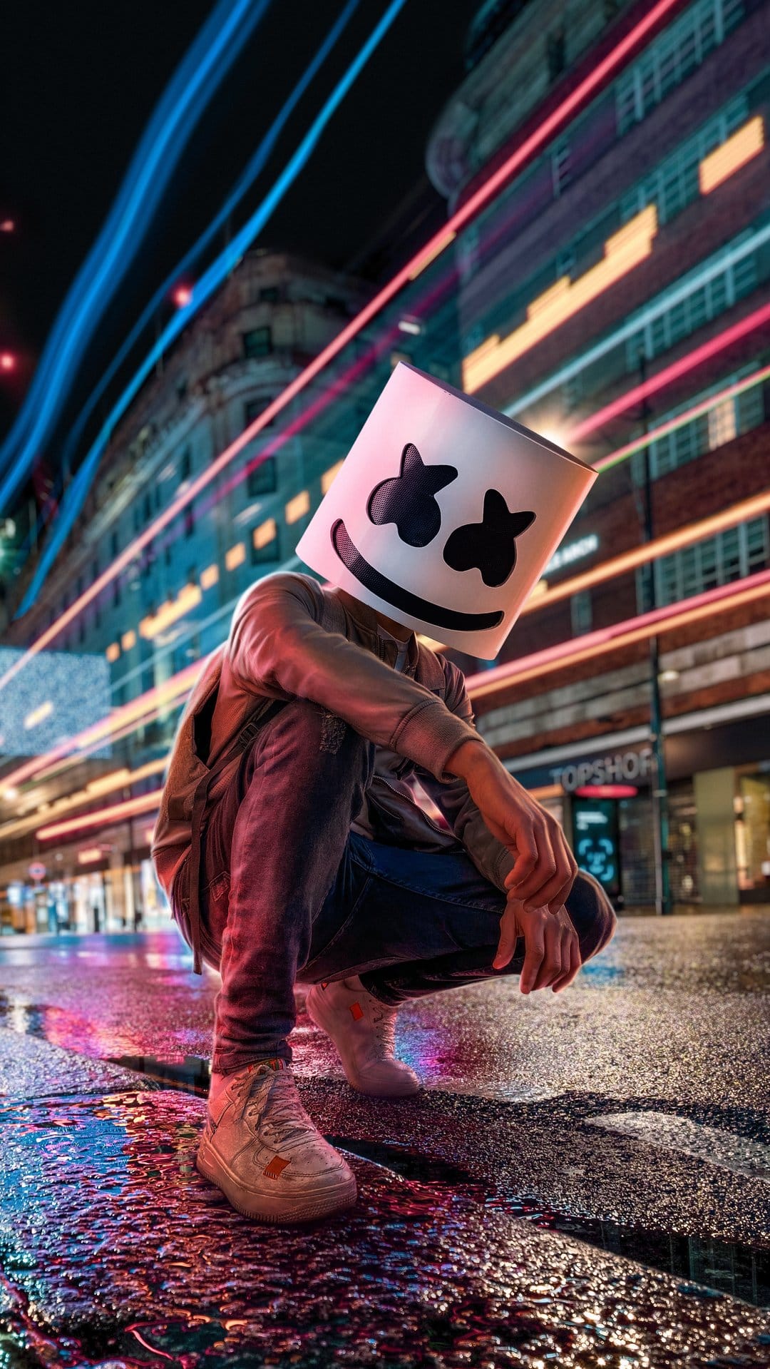 Marshmello-masked figure crouching on a wet asphalt street at night with blurry blue and pink neon light trails.