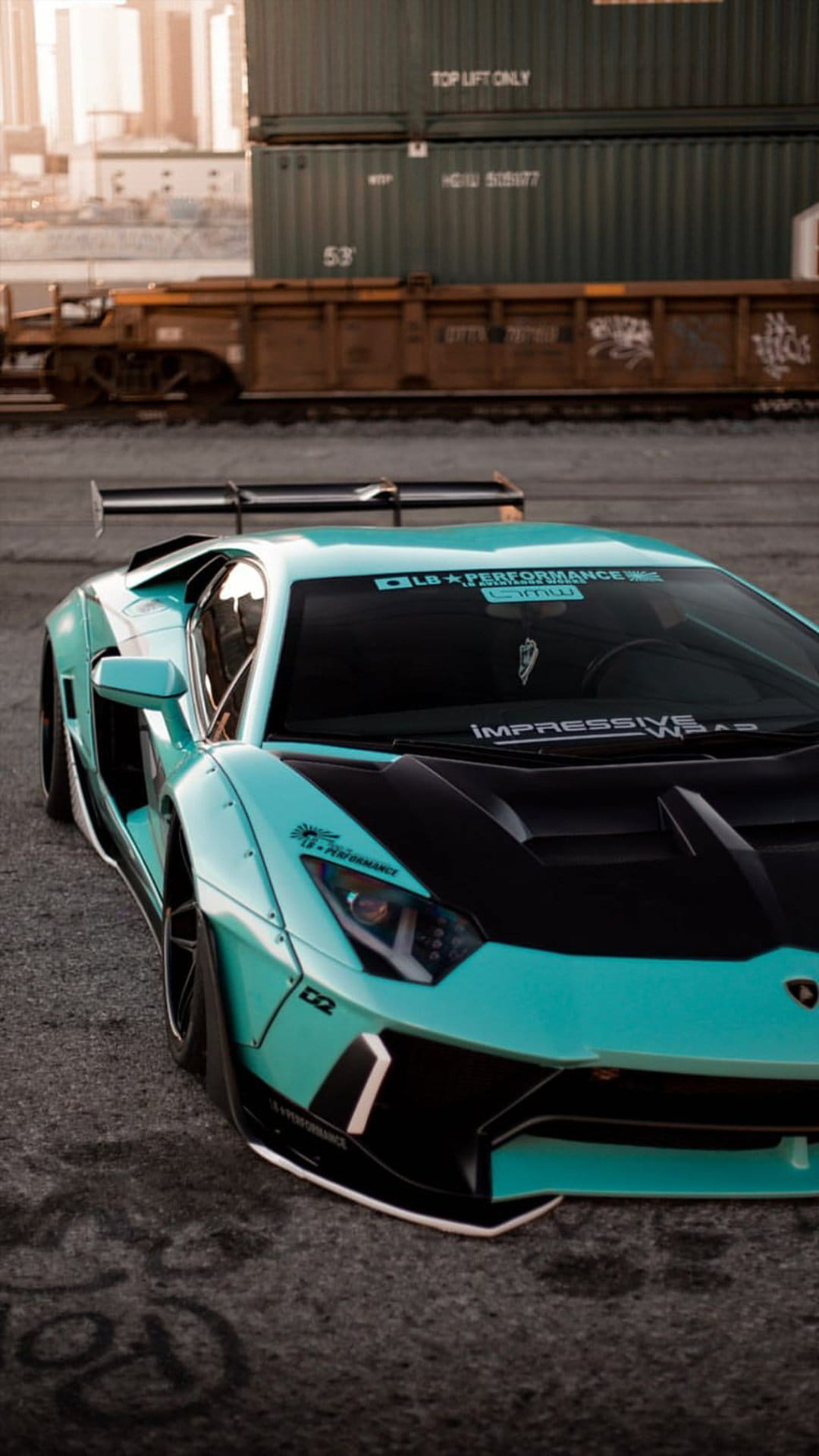 Teal Lamborghini Aventador with a widebody kit parked on asphalt near graffiti and shipping containers.