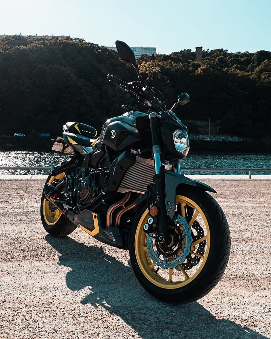Yamaha motorcycle with bright yellow rims parked on gravel near a lake under a clear blue sunny sky and hills.