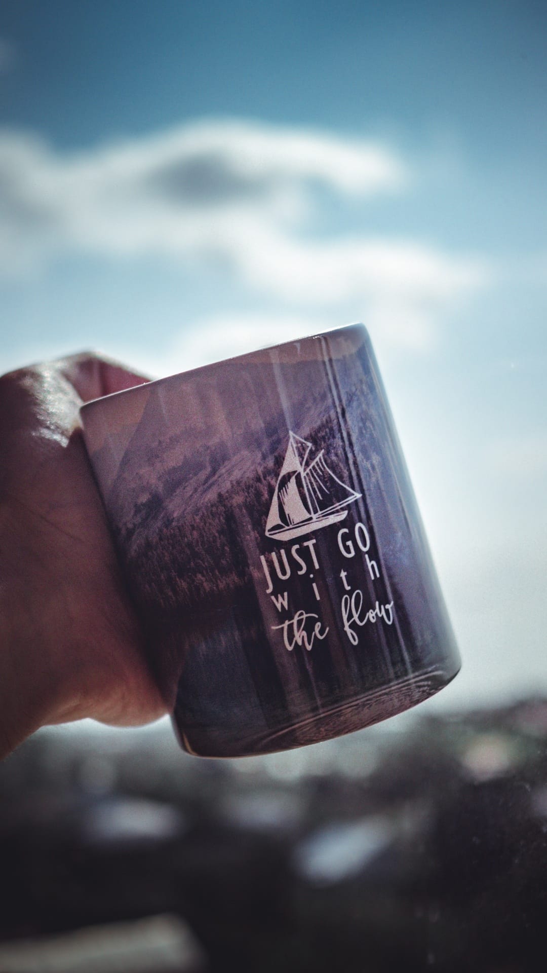 Hand holding a white mug with a sailboat illustration and quote against a bright blue sky with soft clouds.