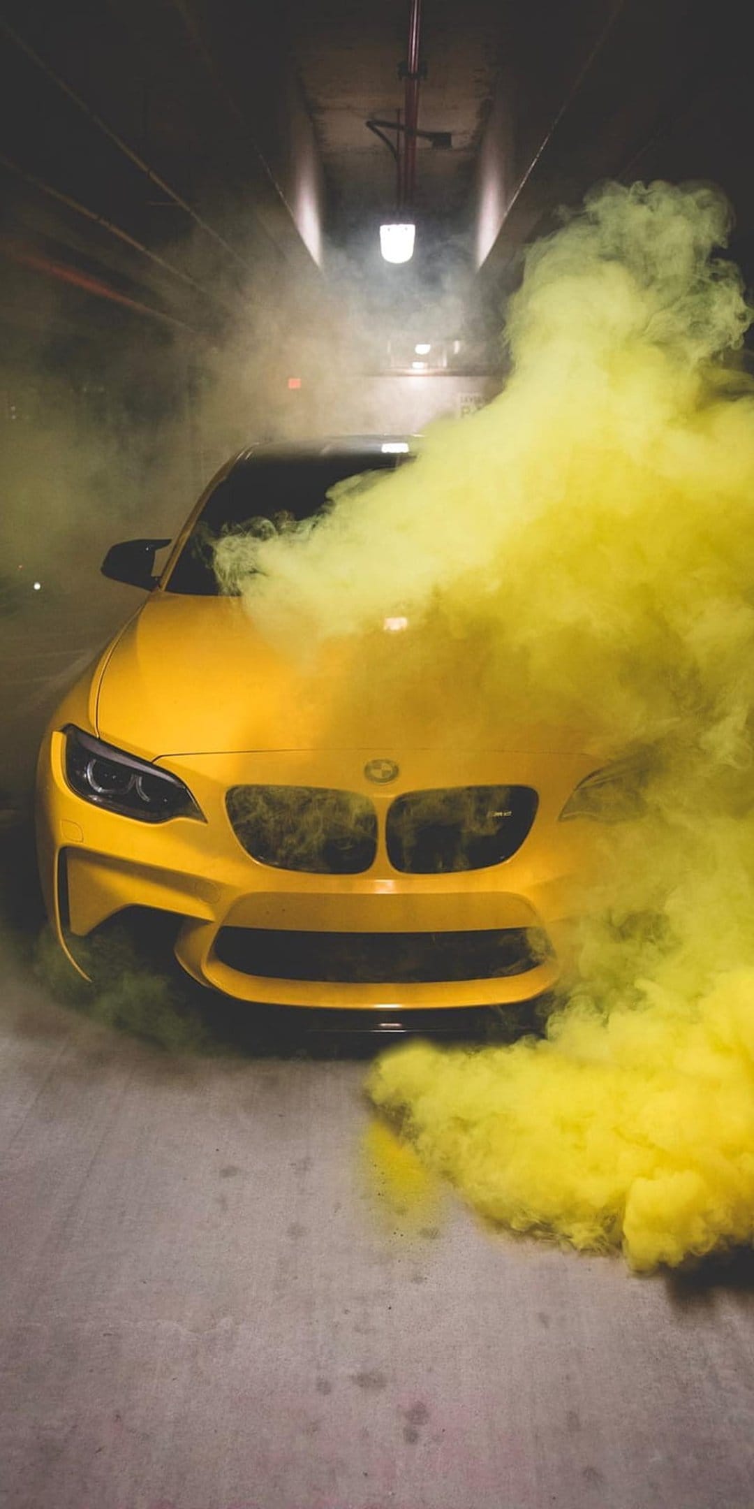 Yellow BMW M2 coupe surrounded by thick yellow smoke in a dark, concrete parking garage with moody lighting.