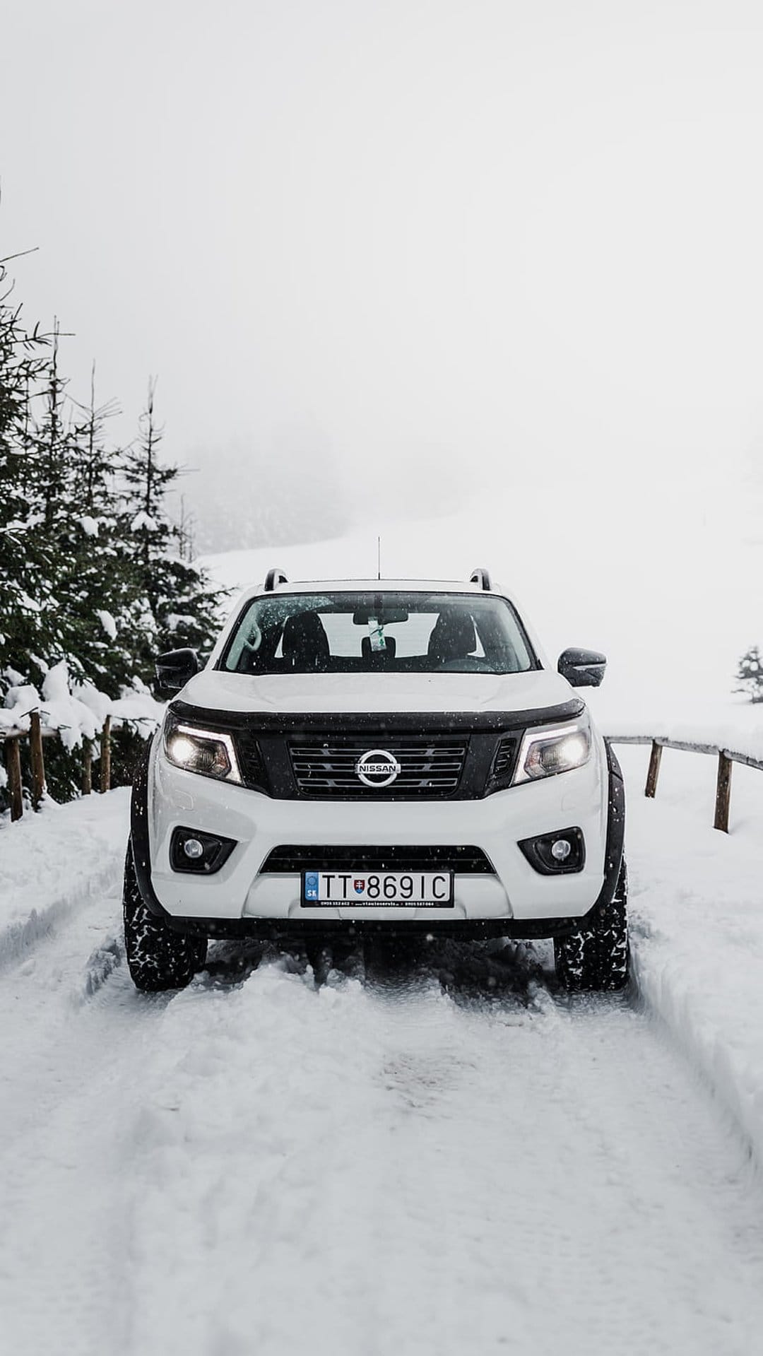 White Nissan Navara pickup truck driving through deep snow on a forest trail with headlights on and pine trees