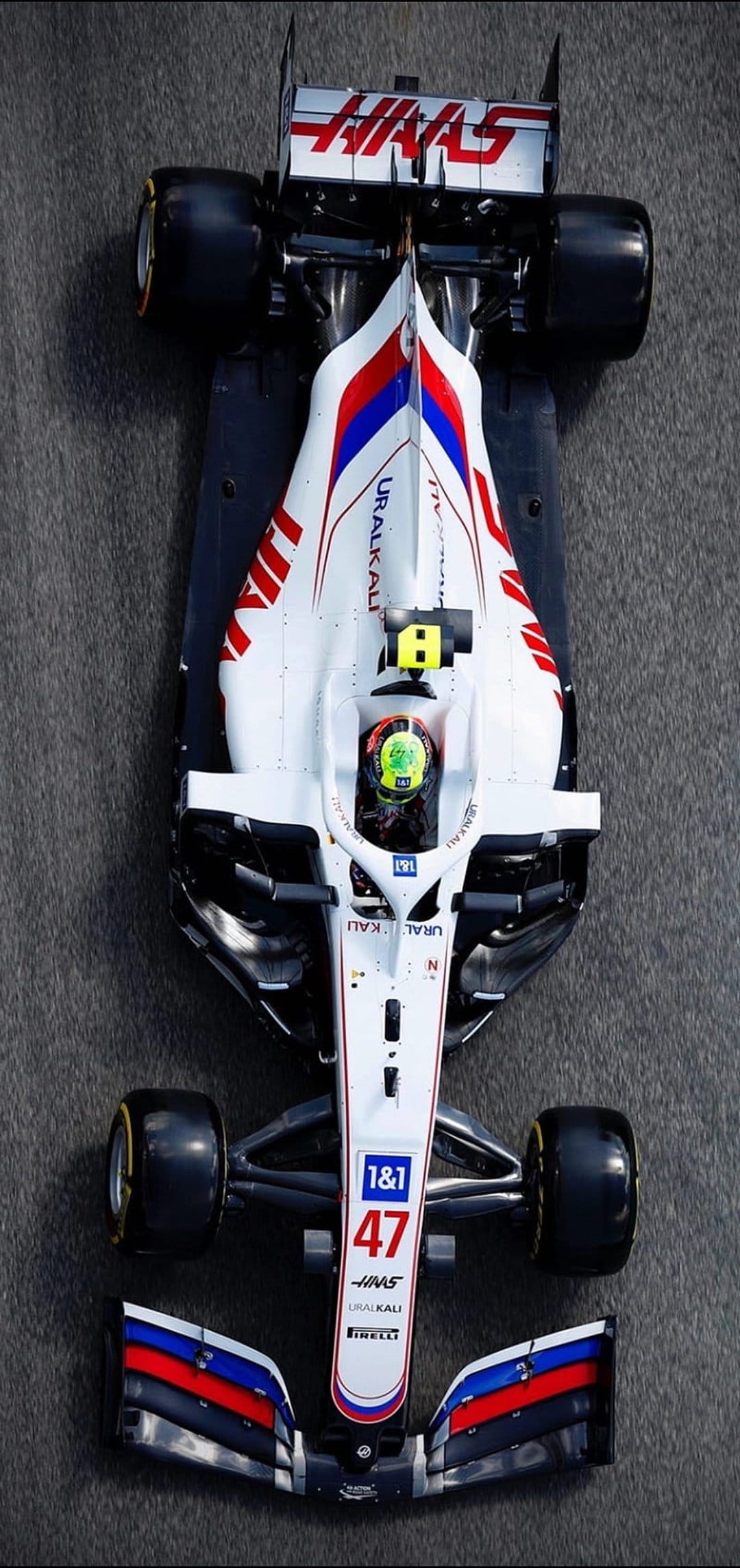 Haas F1 car number 47 with white and red livery seen from a bird's-eye view on a grey asphalt racing circuit.
