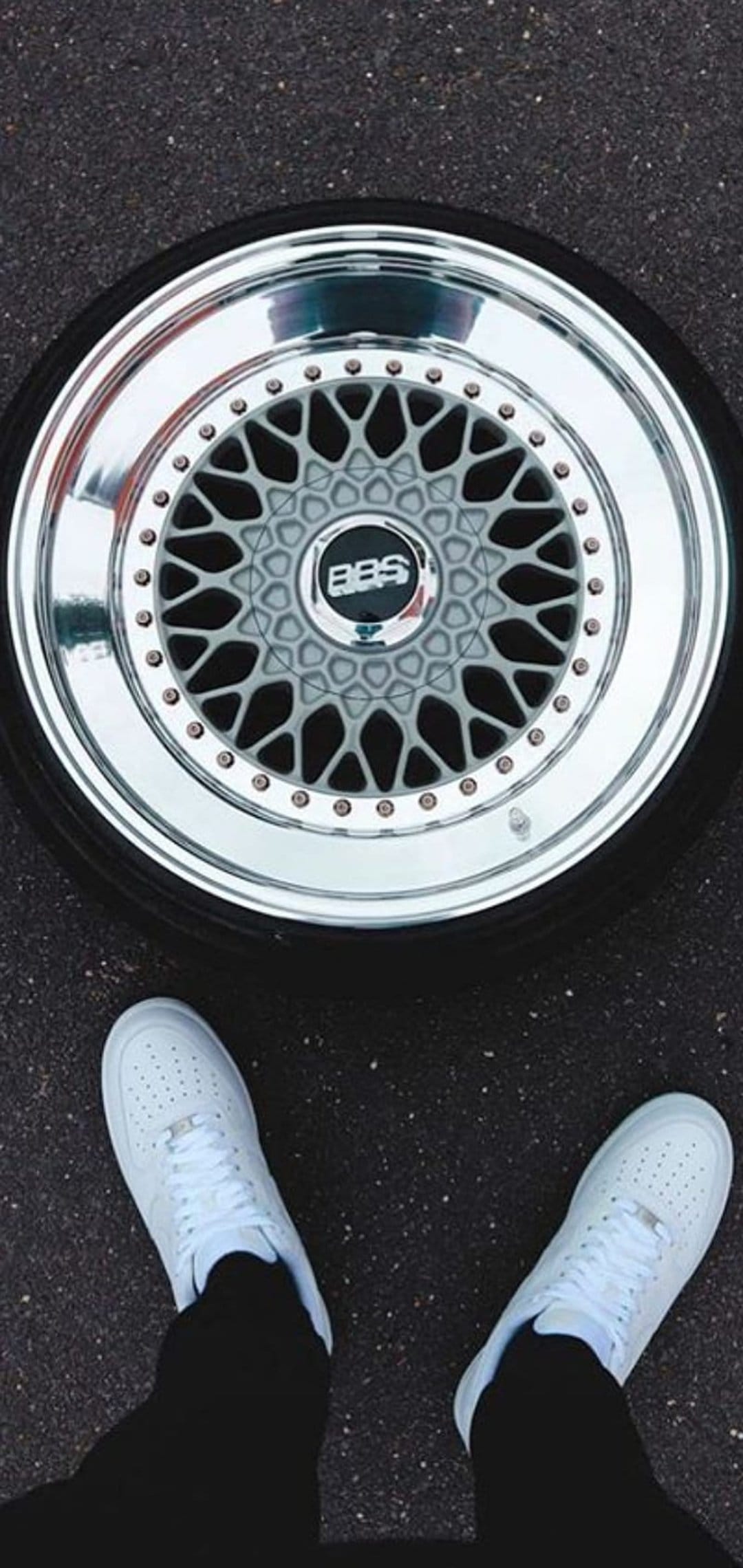 BBS alloy wheel with silver mesh spokes and white Nike sneakers on dark textured asphalt in a vertical layout.