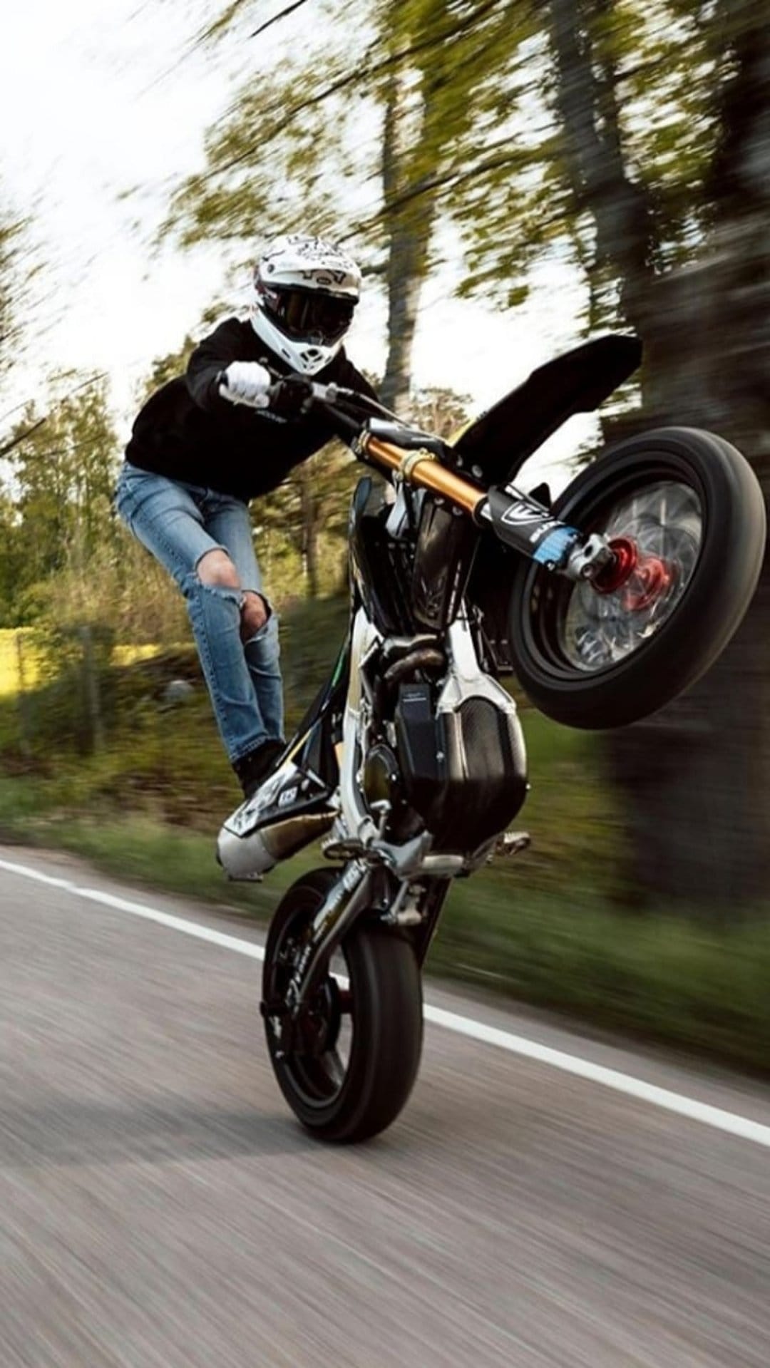 Motorcyclist in a white helmet and black hoodie performs a wheelie on a tree-lined road with motion blur.