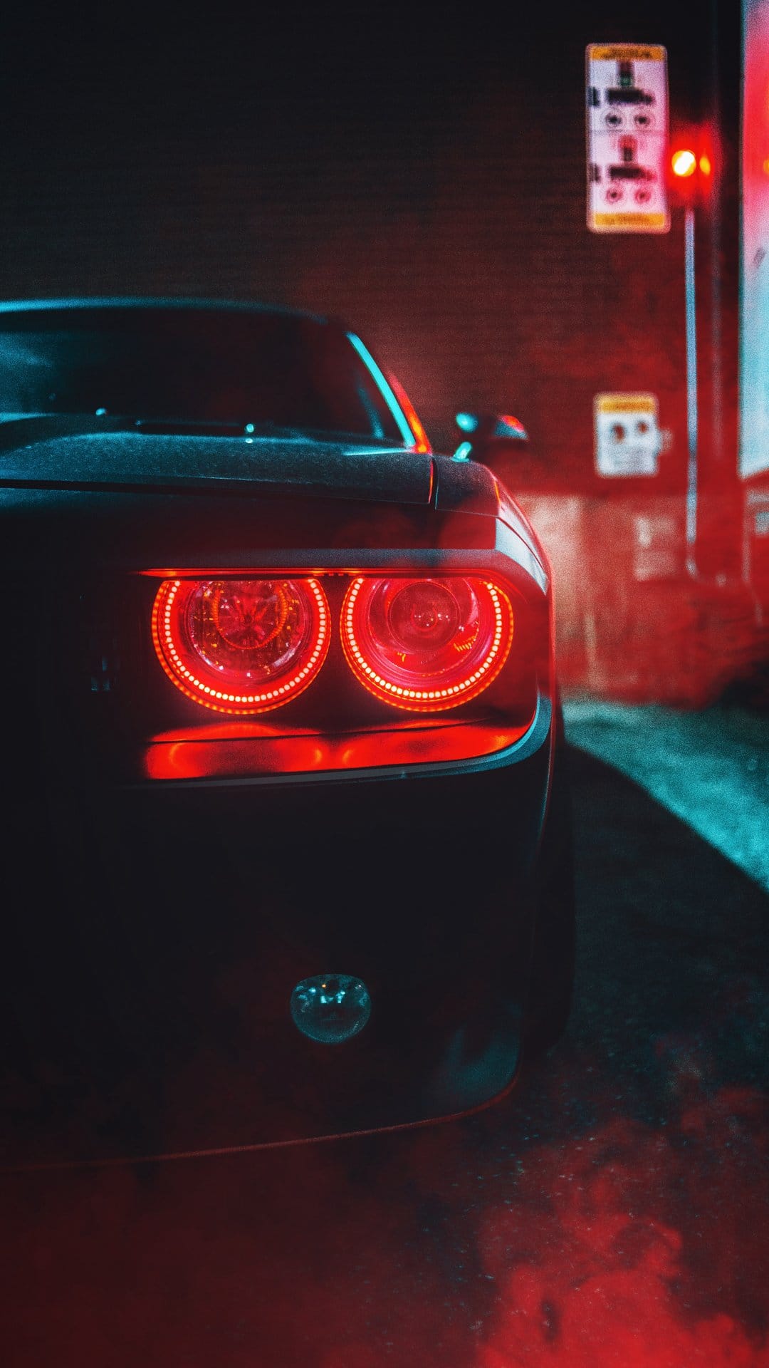 Circular red LED taillights on a dark muscle car reflecting blue neon light against a smoky brick wall at night.