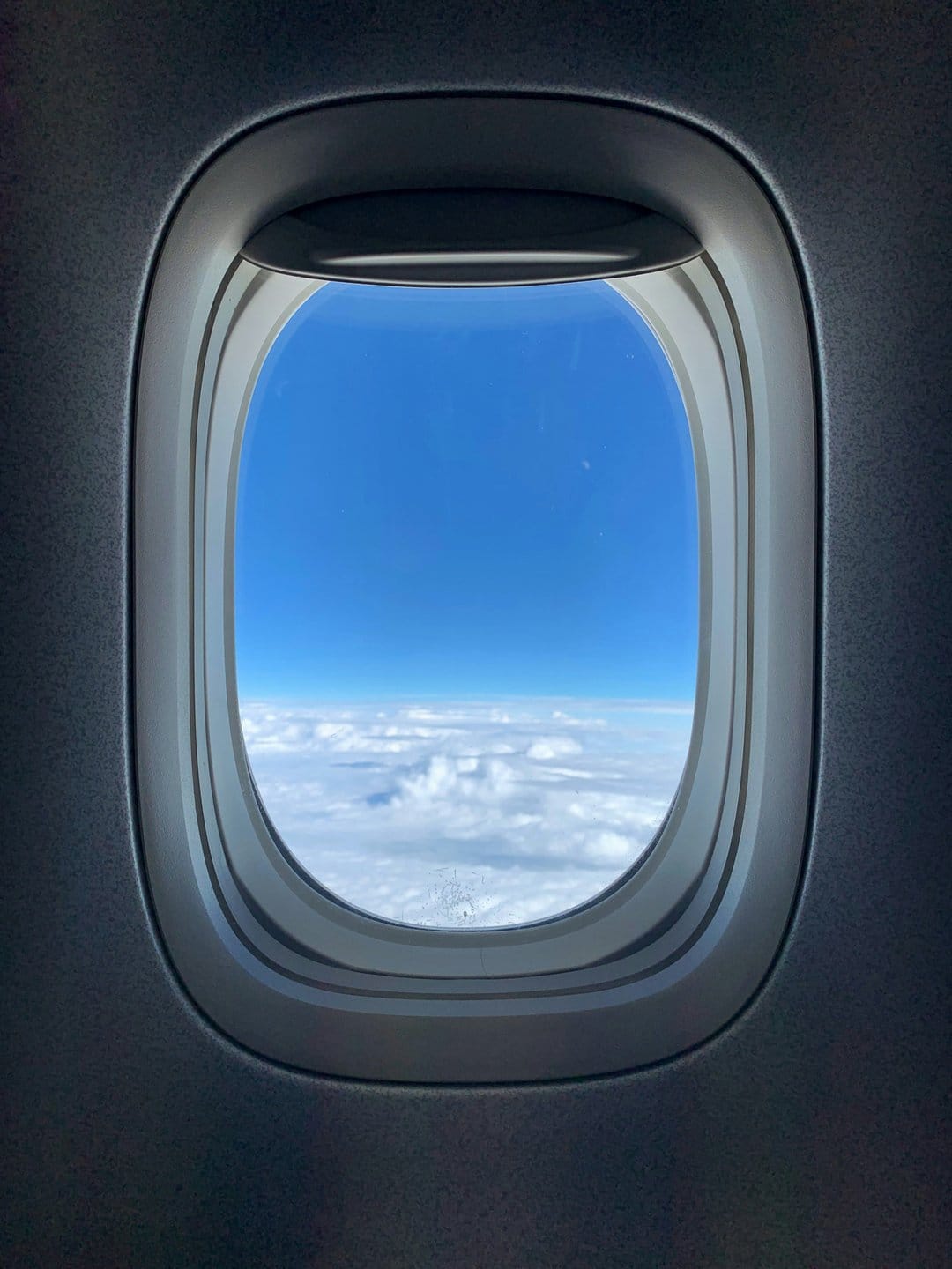 Oval airplane window overlooking a vast expanse of cumulus clouds under a clear blue high-altitude sky.