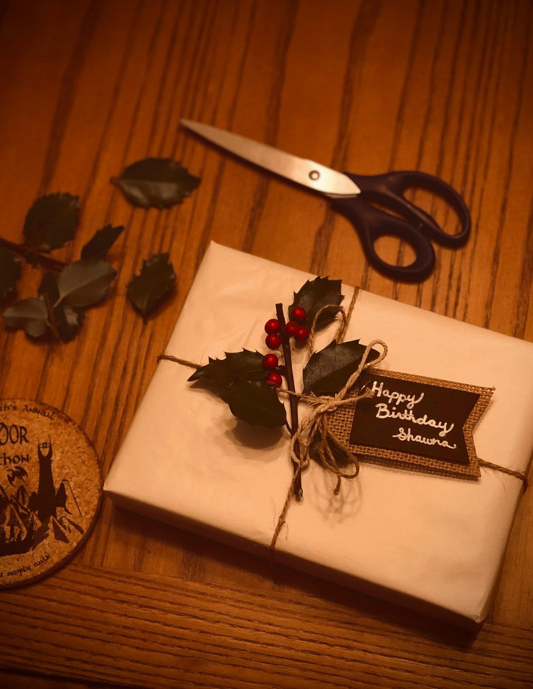 Wrapped birthday gift with a burlap tag, holly sprig, and scissors on a rustic wooden table surface.