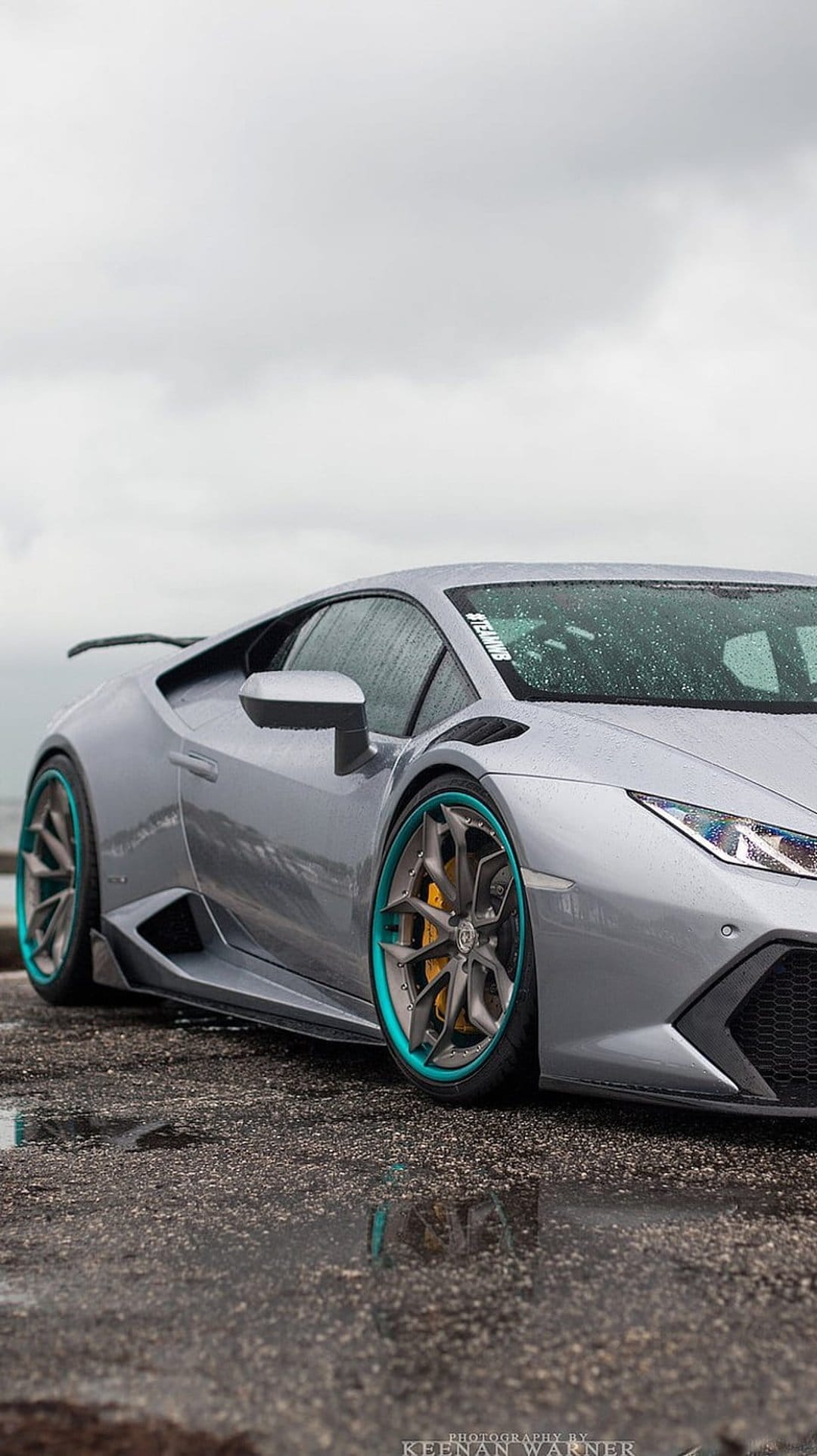 Silver Lamborghini Huracan with teal rims sits on a rainy street with reflections on the wet asphalt surface.