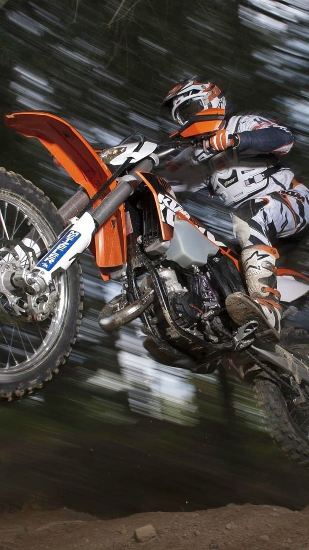 Motocross rider in orange gear jumping a dirt bike mid-air through a blurred forest on a portrait background.