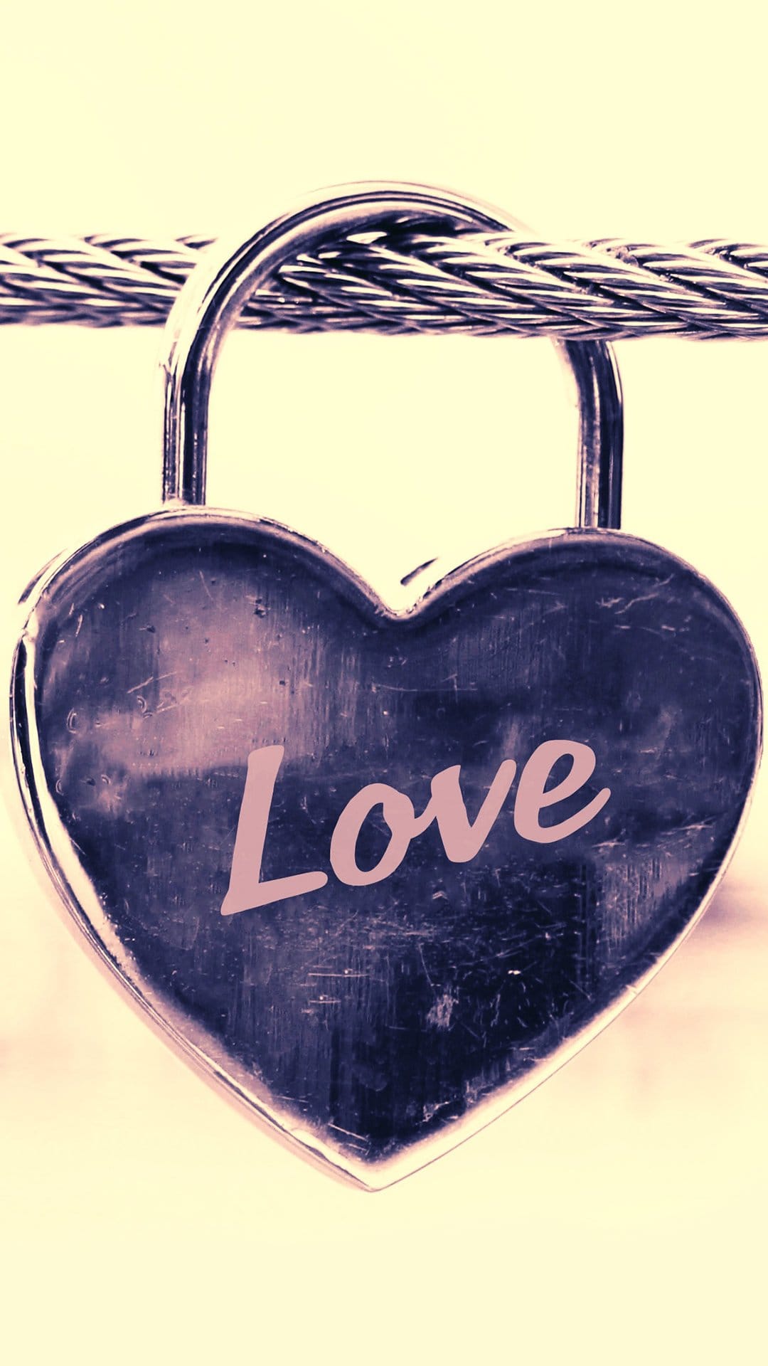 Heart-shaped metal padlock with Love written in pink script, fastened to a thick cable against a yellow backdrop.