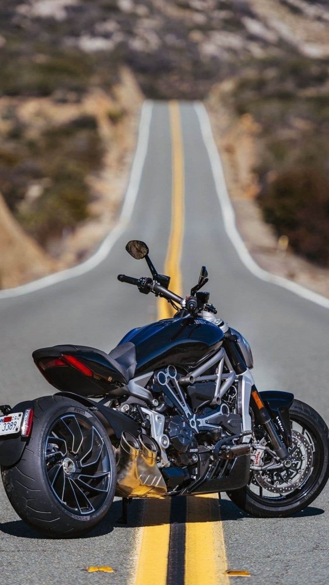 Black Ducati motorcycle parked on an asphalt road with a yellow line stretching through a hilly desert landscape.