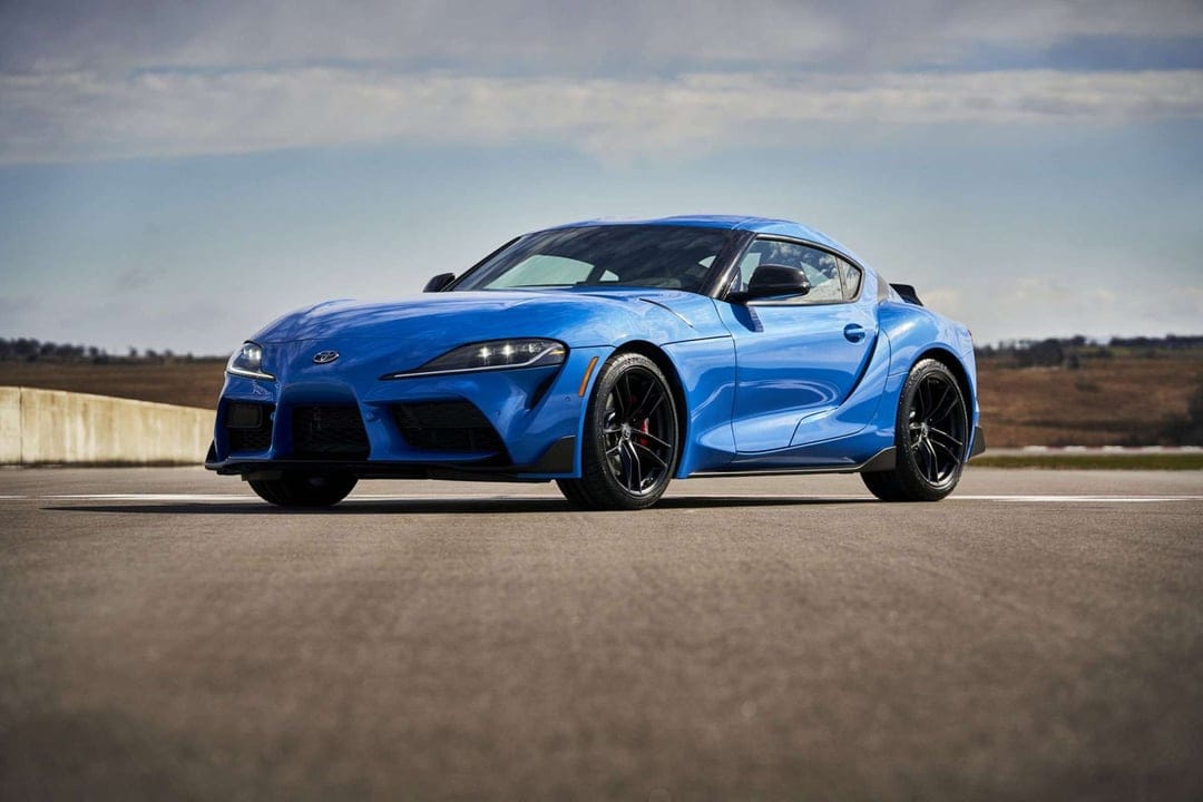 Vibrant blue Toyota GR Supra sports car with black wheels parked on a racetrack under a bright overcast sky.