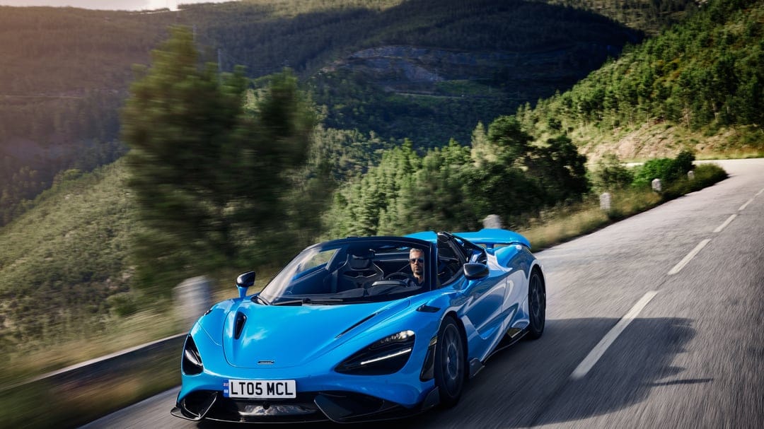 Blue McLaren 765LT Spider supercar speeding on a winding mountain road with motion-blurred green forest background.