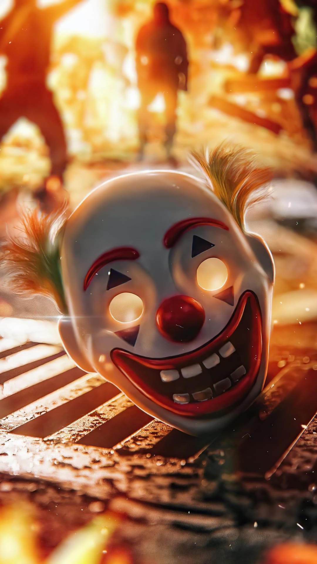 White clown mask with glowing yellow eyes and orange hair rests on a metal grate against a blurry fire.