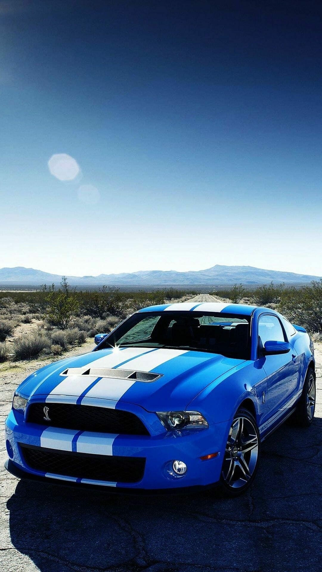 Blue Shelby GT500 with white racing stripes on a desert road under a clear sky with lens flare.