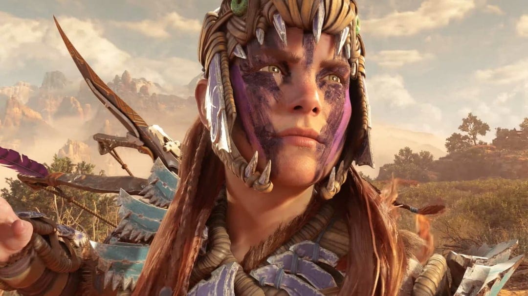 Aloy with purple war paint and a bone headdress stands against a sunlit mountain landscape with her bow.