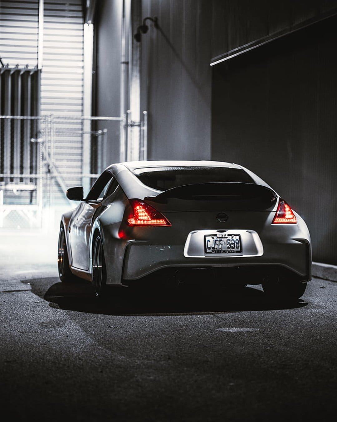 Nissan 370Z rear end parked in a dark alley with glowing red taillights and a visible license plate on silver paint.