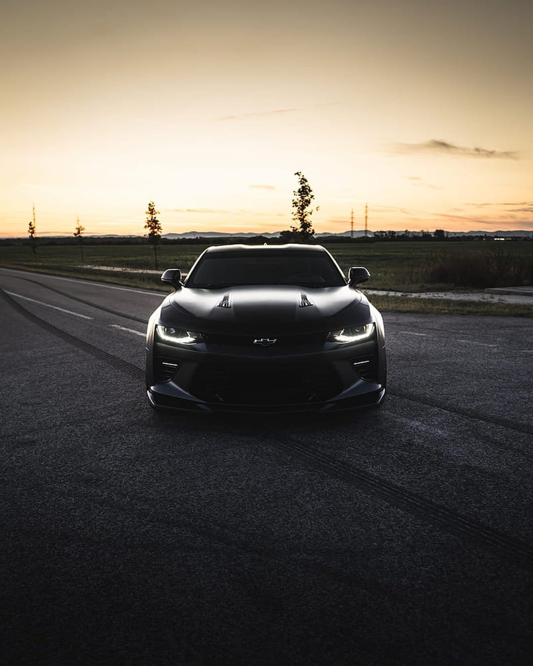Chevrolet Camaro with lit headlights rests on a dark road against a warm twilight horizon and silhouettes.