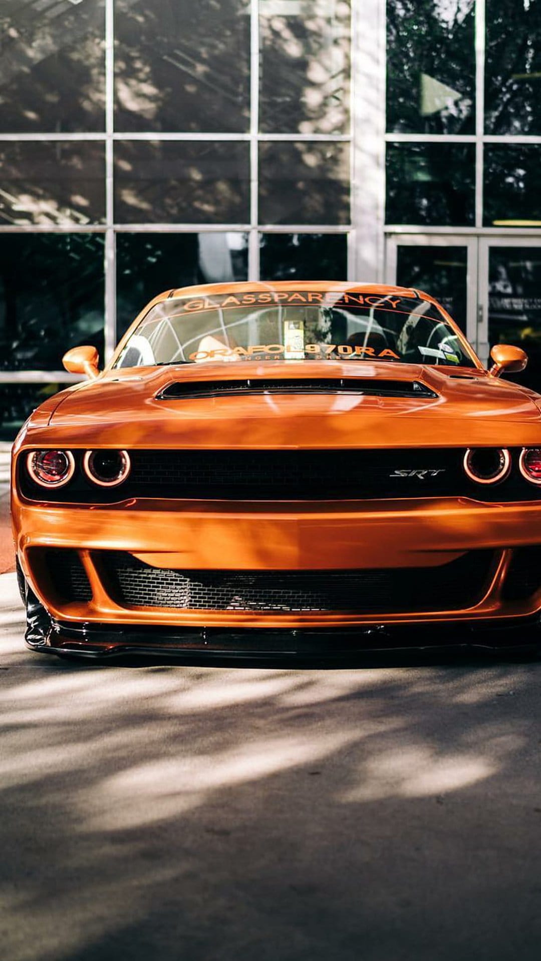 Orange Dodge Challenger SRT Hellcat with glowing halo headlights parked on asphalt before a glass office building.