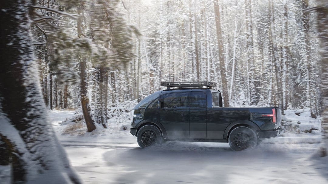 Black electric pickup truck driving on a snowy woodland road surrounded by frost-covered evergreen trees.