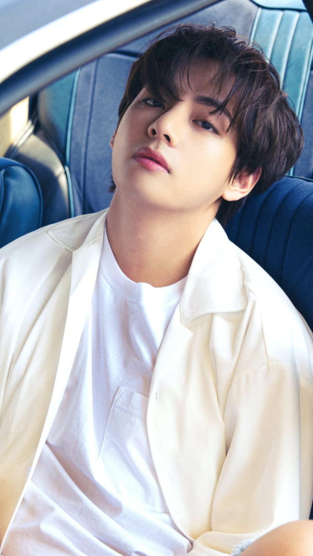Kim Taehyung of BTS tilts his head back in a car, wearing a white shirt with a soft-focus background.