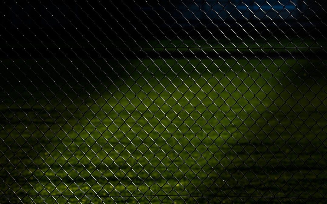 Dark chain-link fence with a diamond pattern in front of an artificially lit green sports field at night.