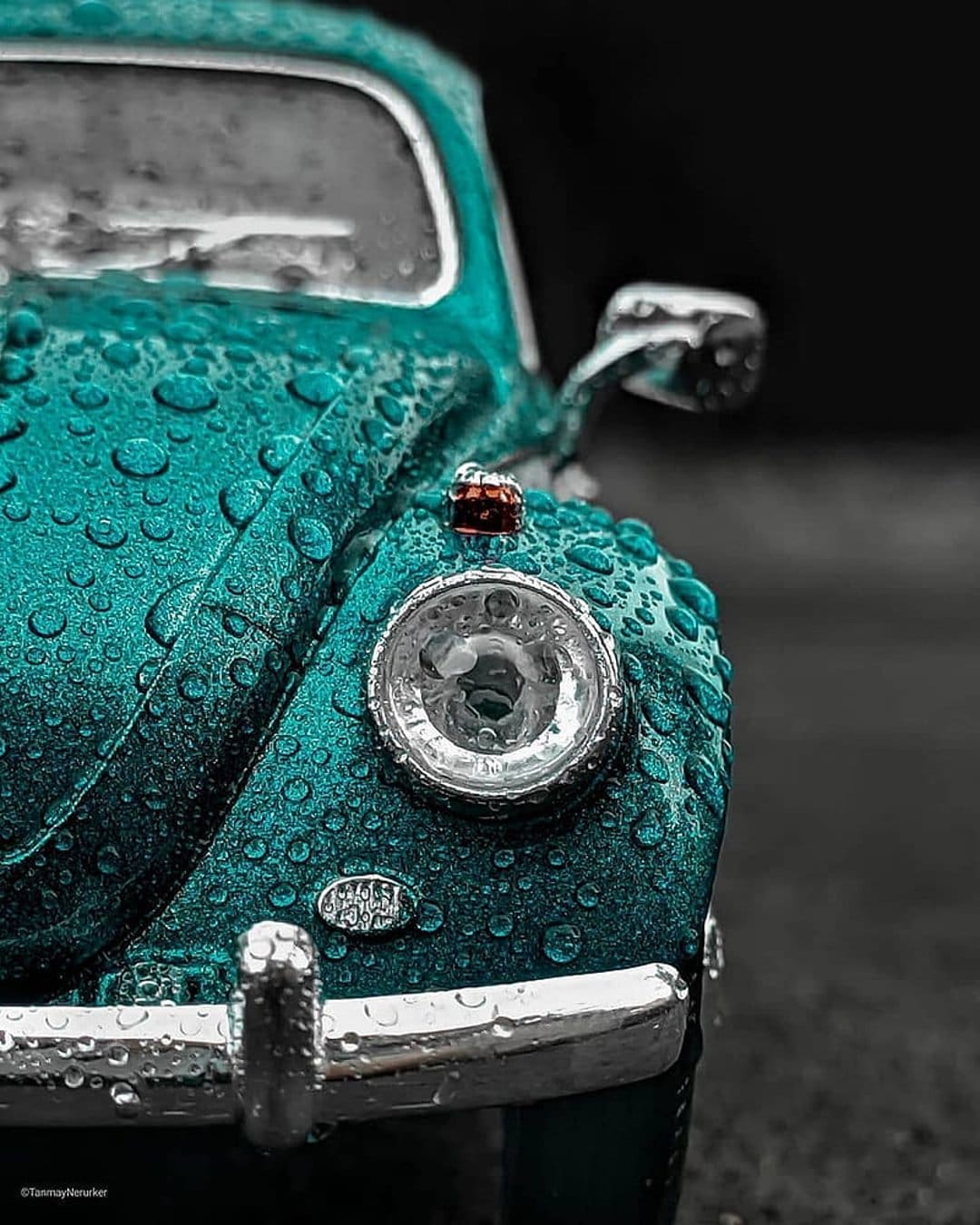 Teal Volkswagen Beetle headlight and chrome bumper covered in small water droplets with a blurred dark background.