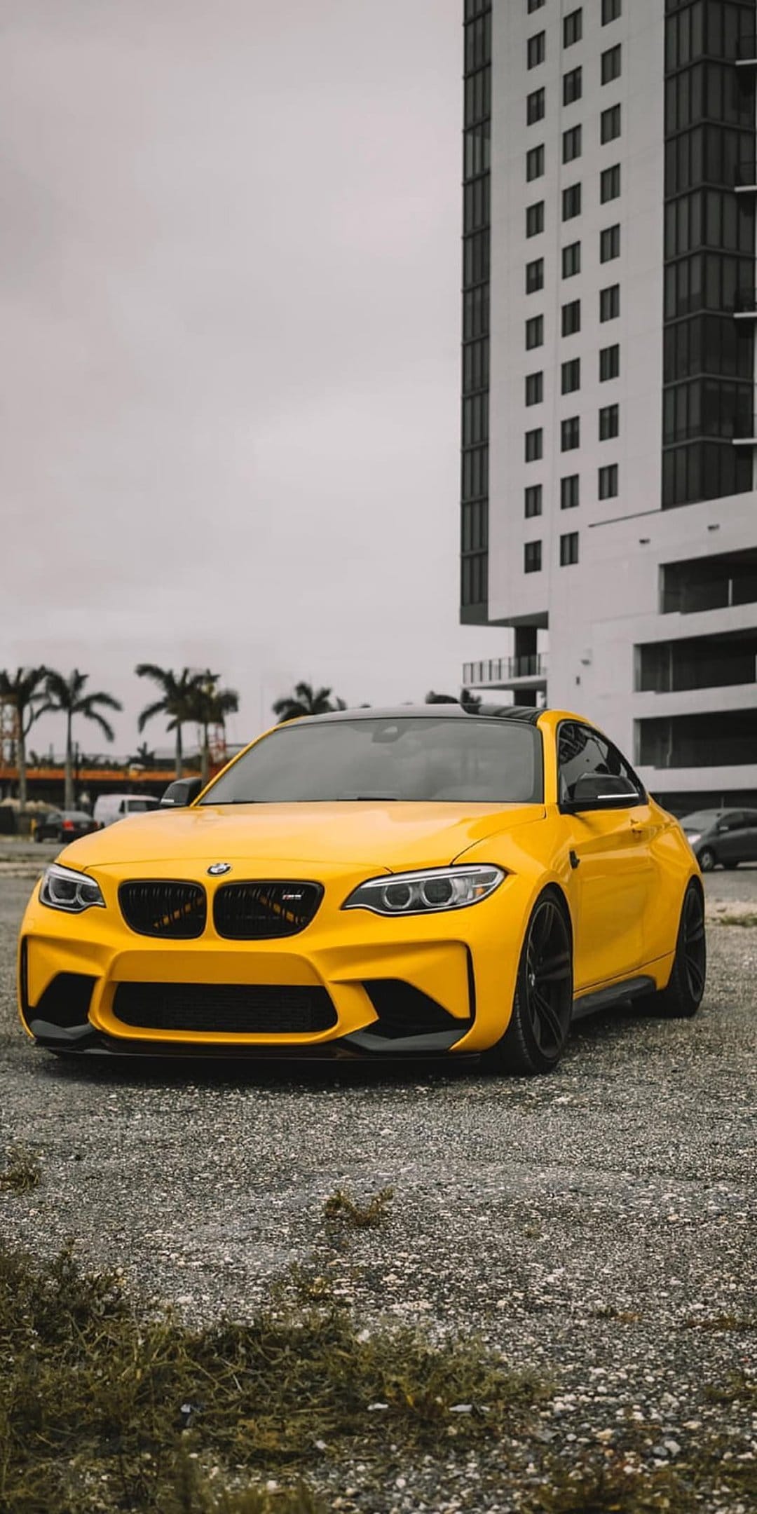 Yellow BMW M2 parked on gravel with a tall white modern building and green grass under a gray overcast sky.