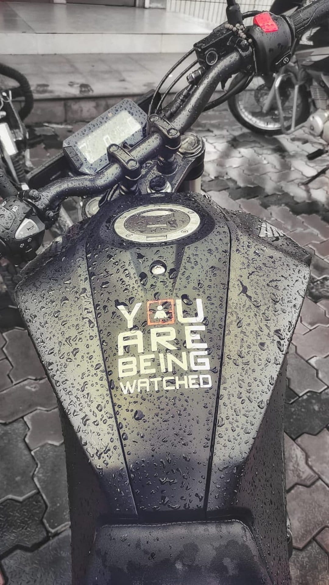 Black motorcycle fuel tank covered in raindrops with a white You Are Being Watched decal and orange icon.